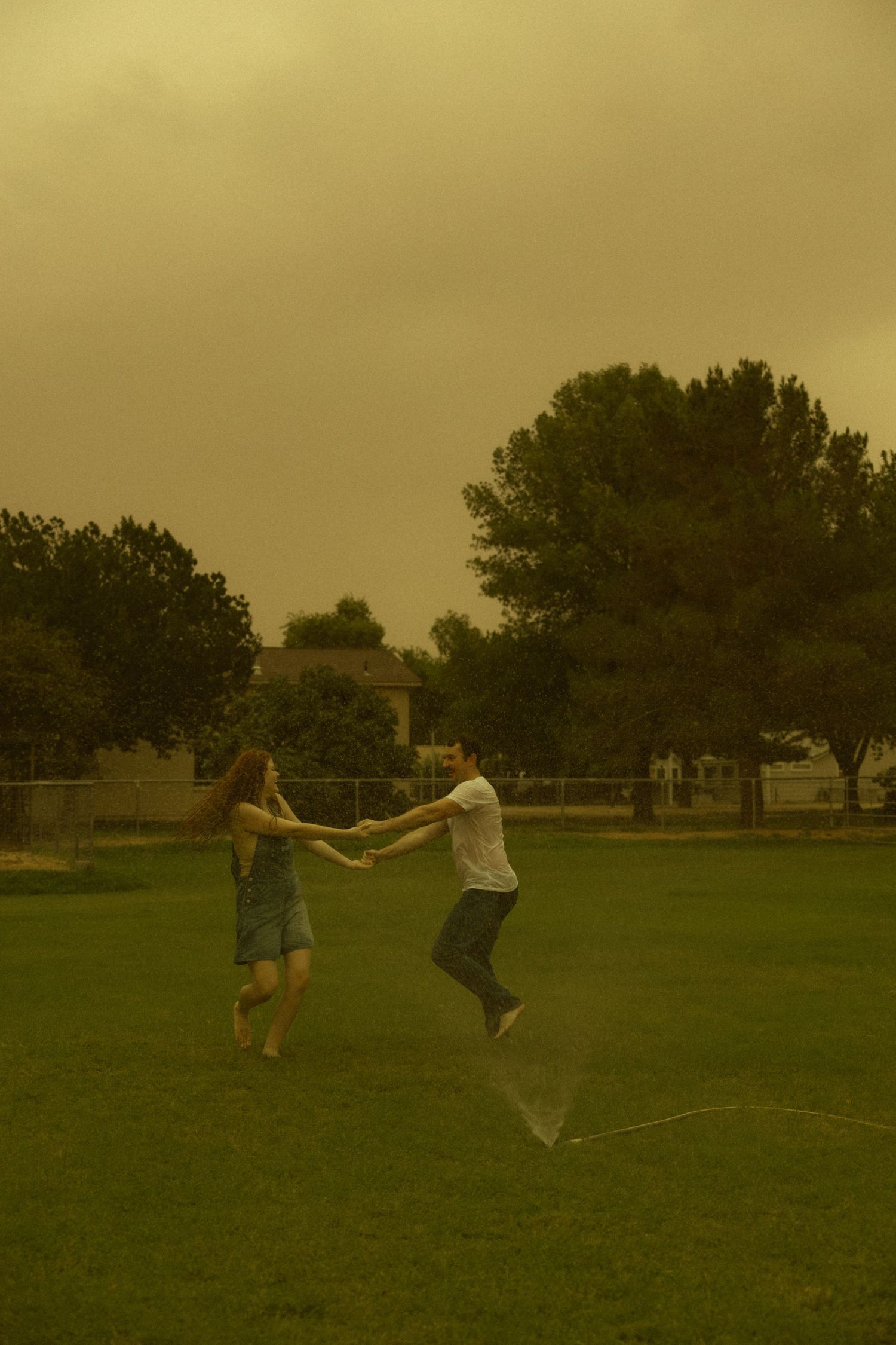Couple holding hands and jumping in a green park under a dramatic orange-tinted stormy sky.