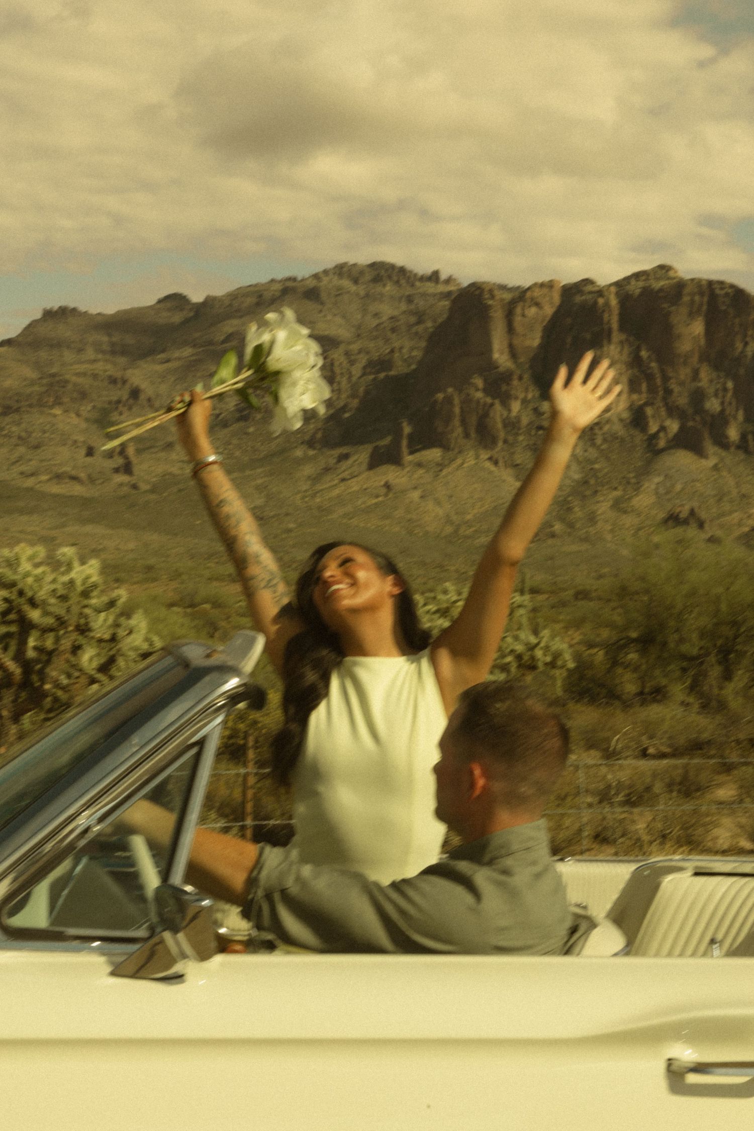 A joyful bride raises her arms with a bouquet in a white convertible, with a man beside her and desert mountains behind.