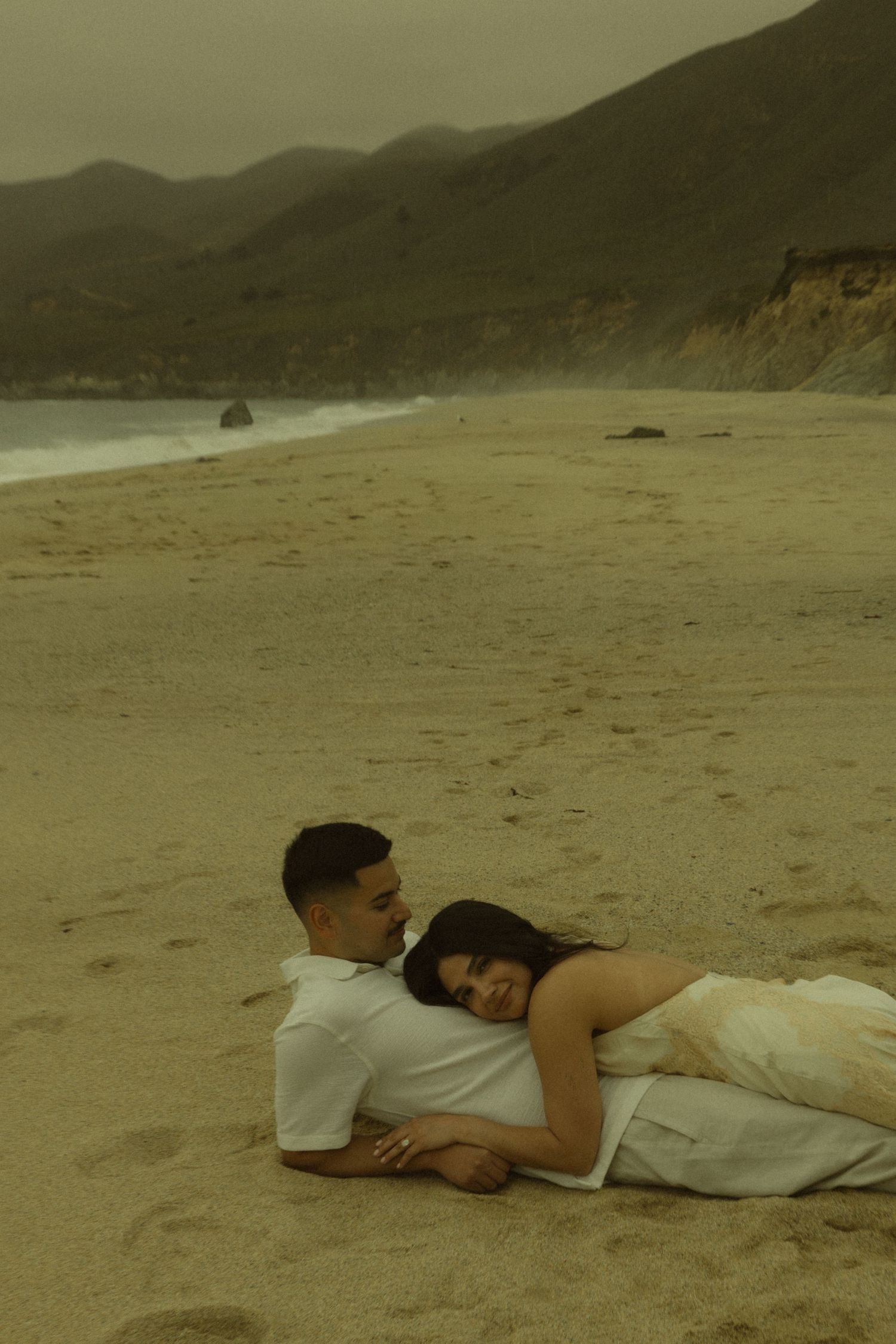 Romantic couple cuddling on beach sand with mountain cliffs and cloudy sky in background.