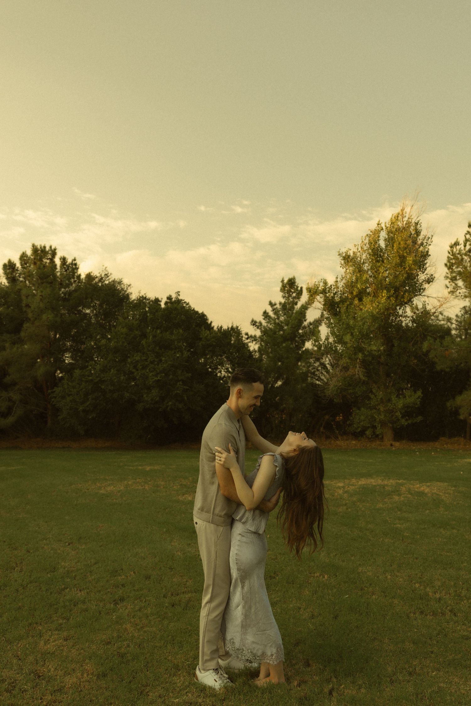 Romantic couple embracing in a lush green park at golden hour, surrounded by trees under a soft evening sky.