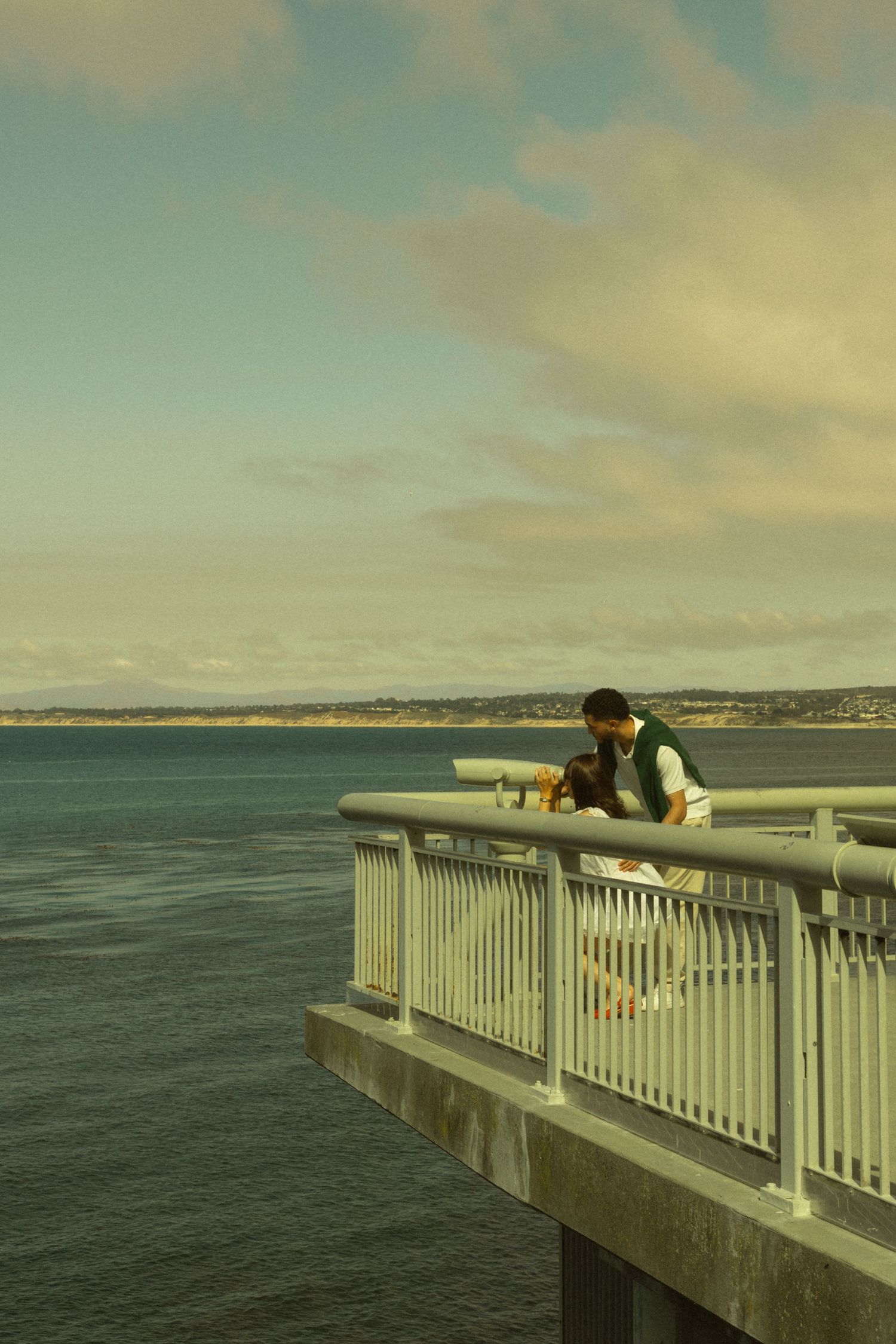 Couple sharing a romantic moment on a seaside pier overlook with ocean views and cloudy sky backdrop.