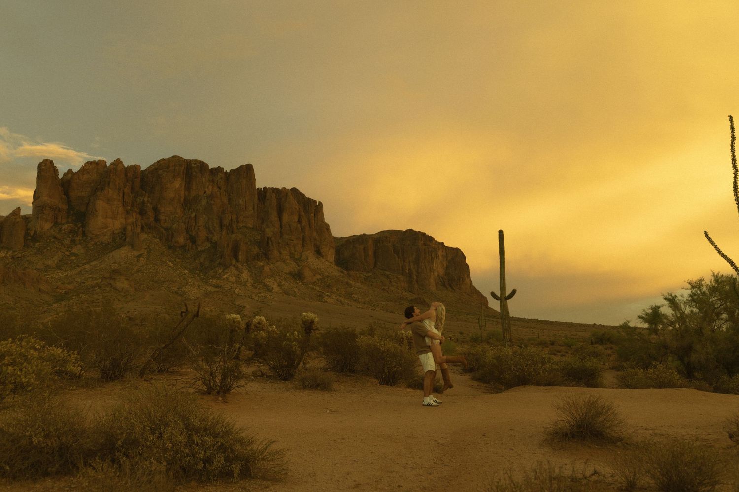 Couple embracing in Arizona desert at sunset with Superstition Mountains and saguaro cactus in golden light.