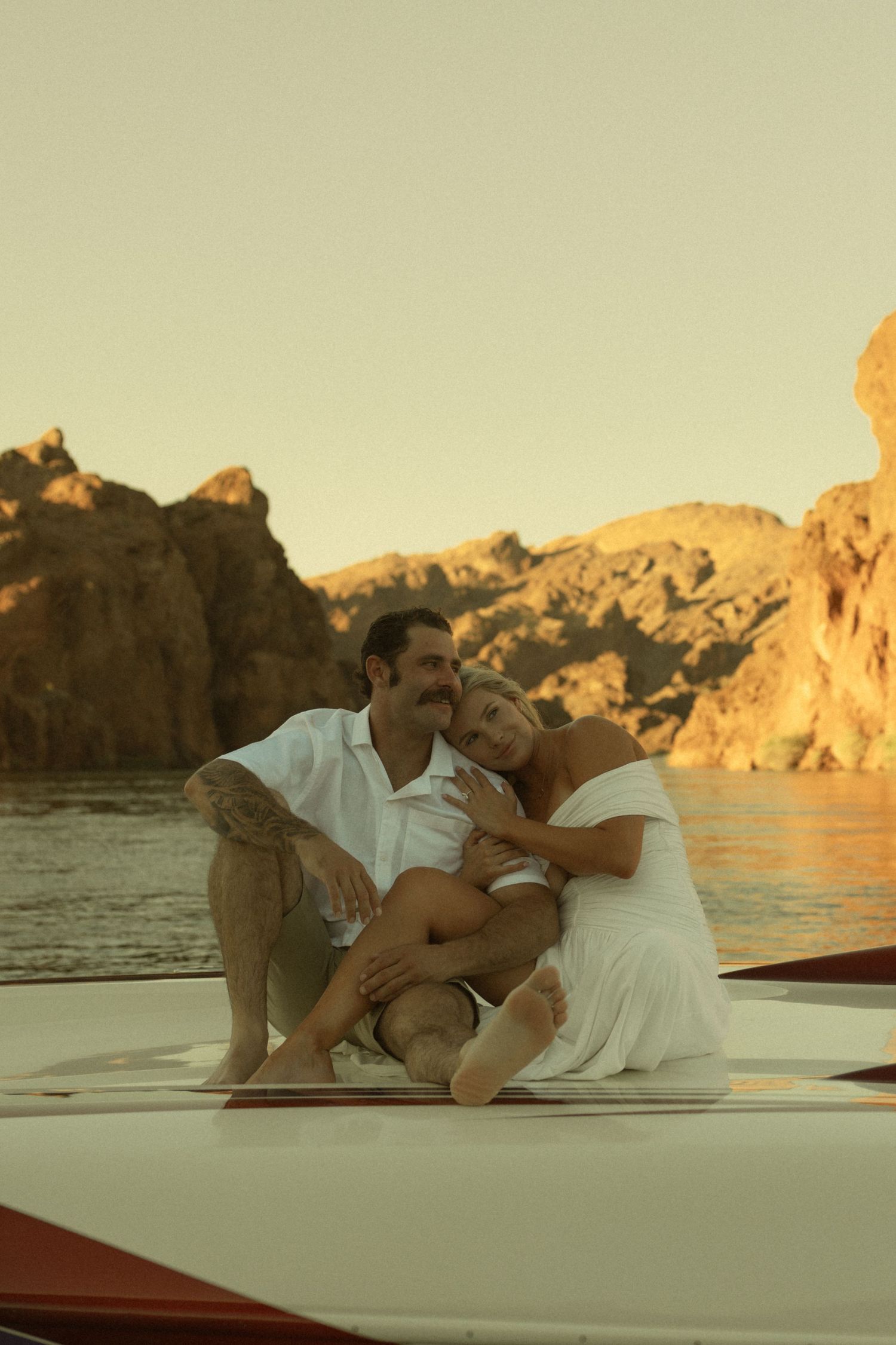 Couple sitting together on a boat at sunset with rocky cliffs and calm water in the background.