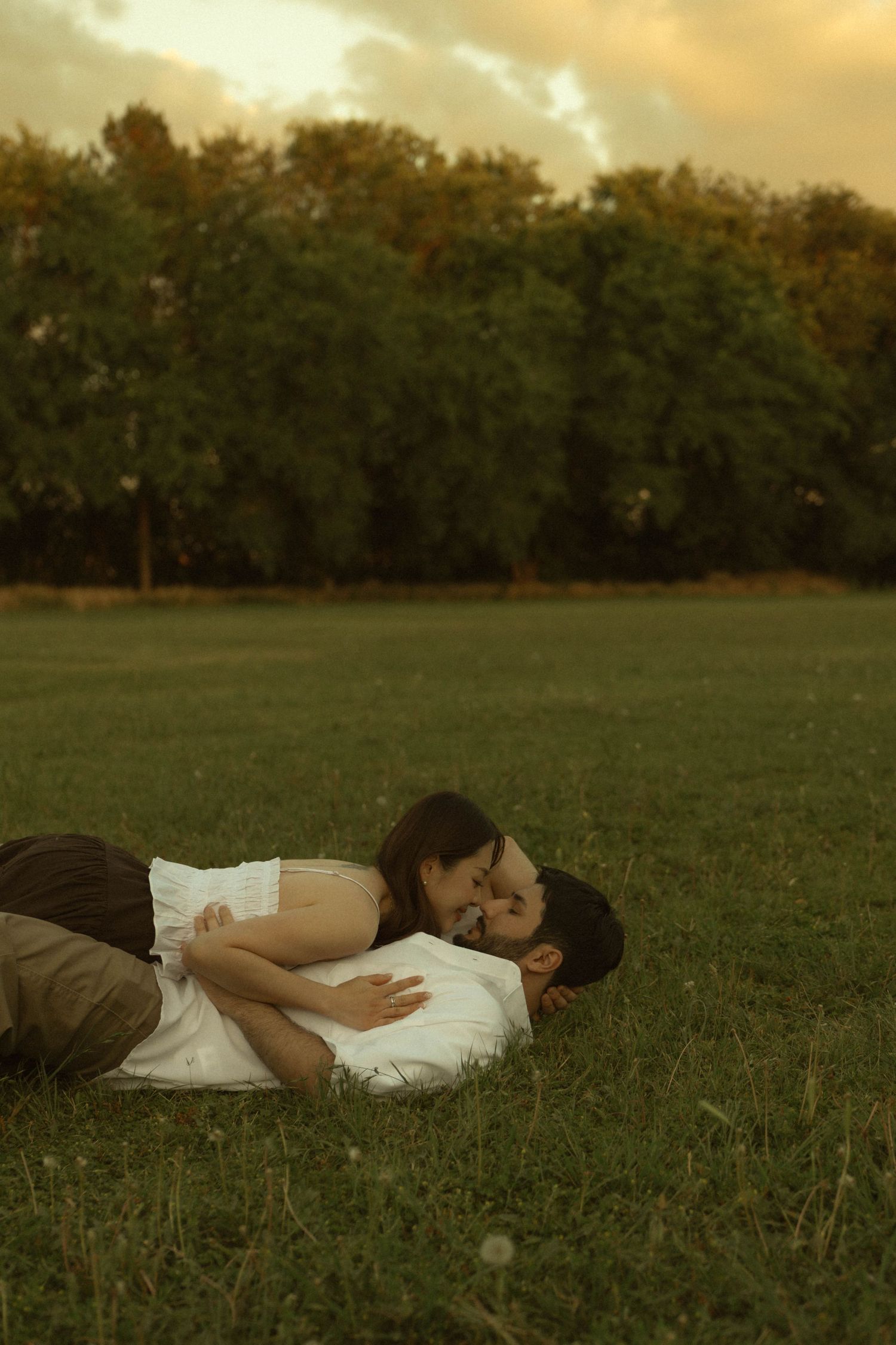Romantic couple lying on green grass in a park at sunset, embracing tenderly with trees in the background.