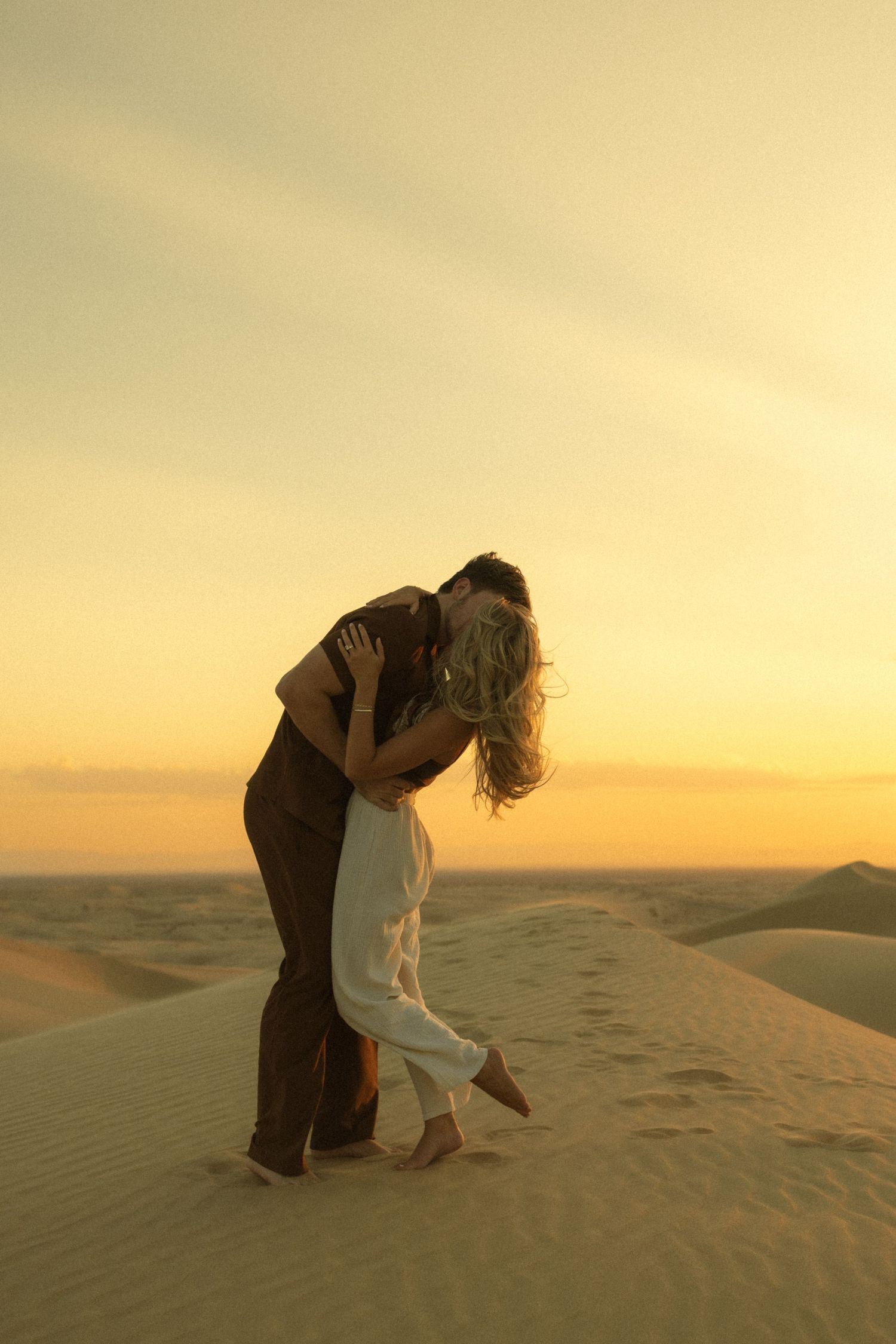 Romantic couple kissing on a sand dune during a warm desert sunset.