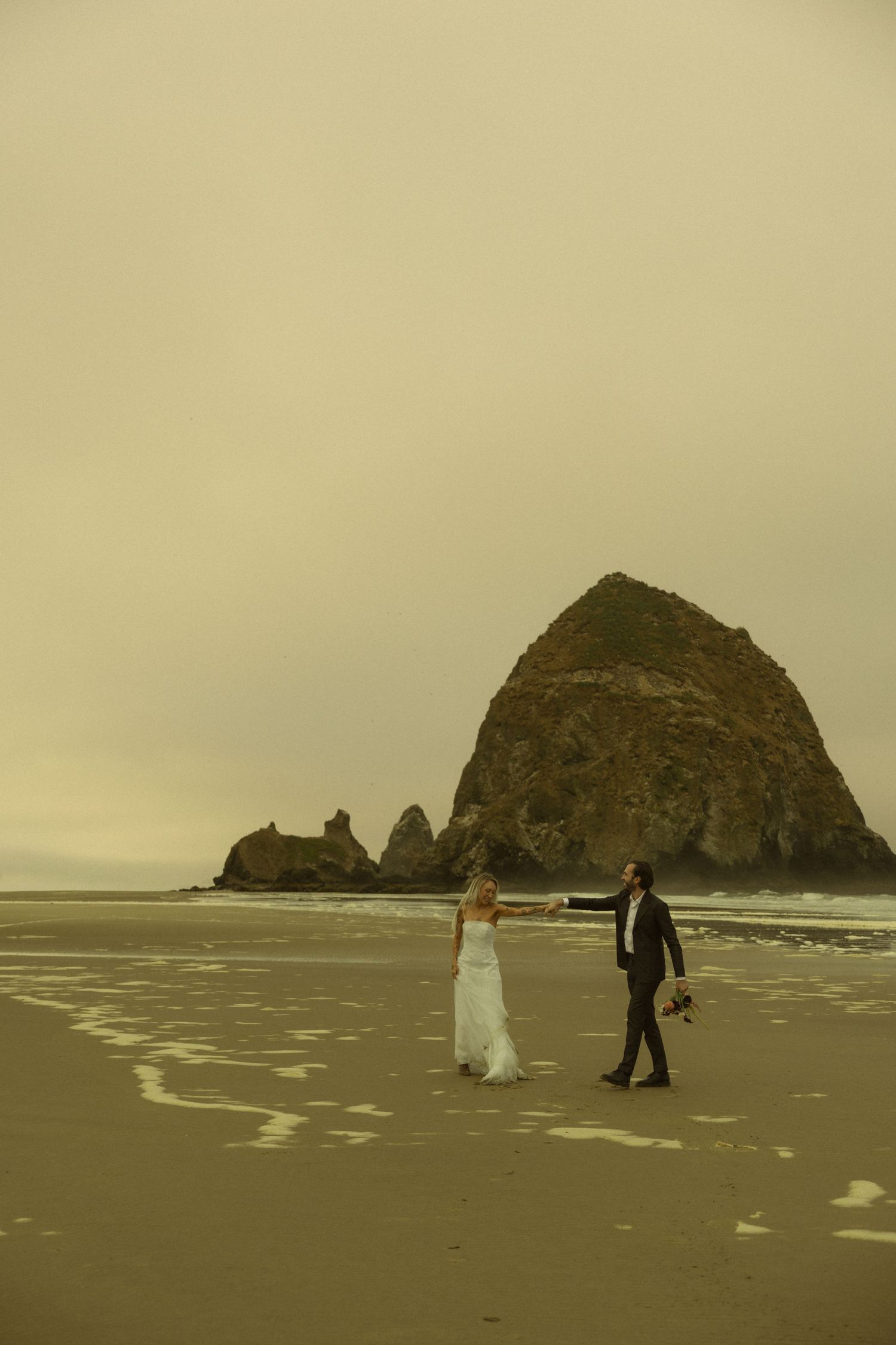 Wedding couple walks hand in hand on sandy beach with iconic Haystack Rock at Cannon Beach, Oregon.
