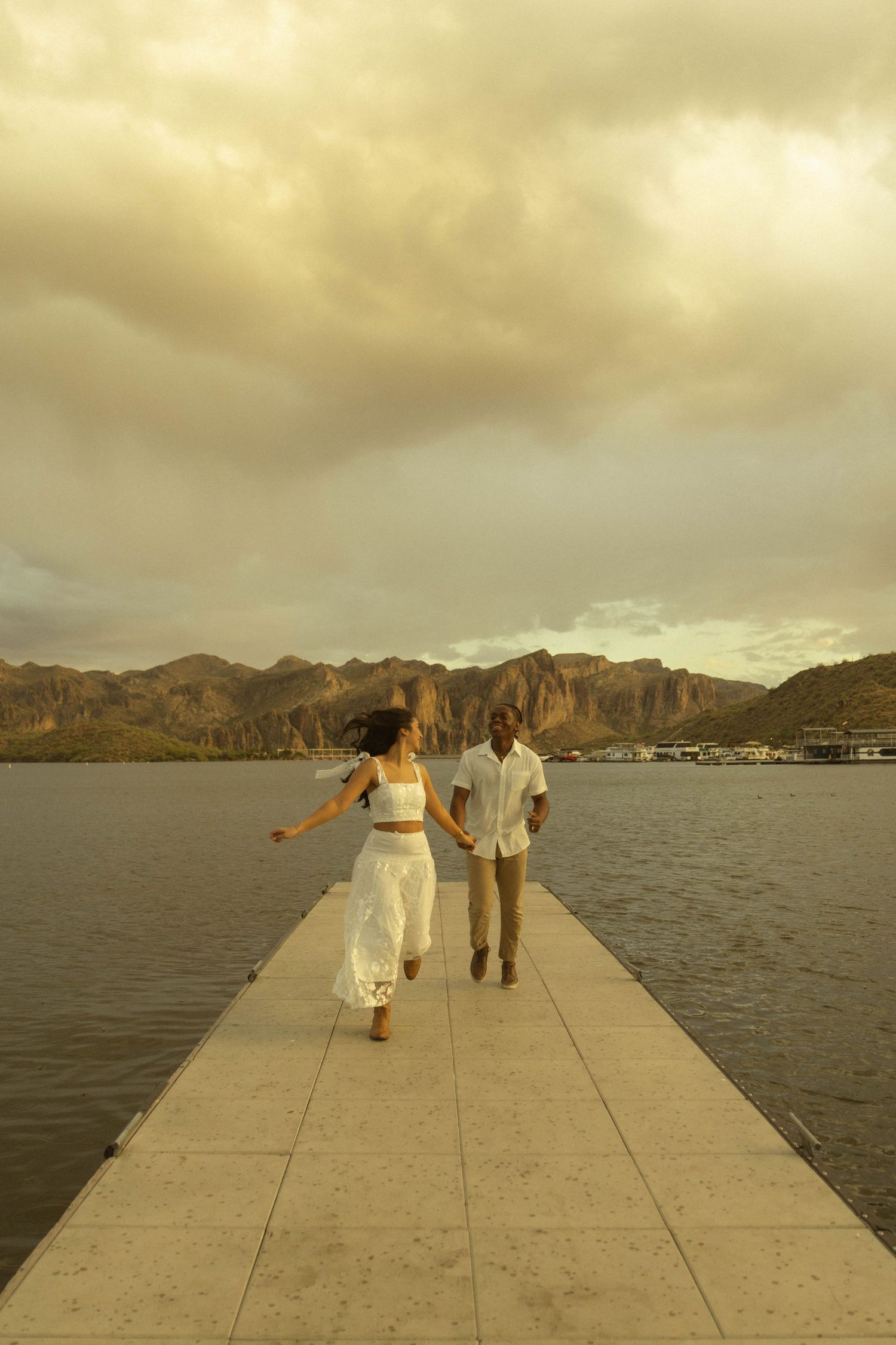 Couple walking hand in hand down a scenic lakeside pier with mountains and cloudy sunset sky.