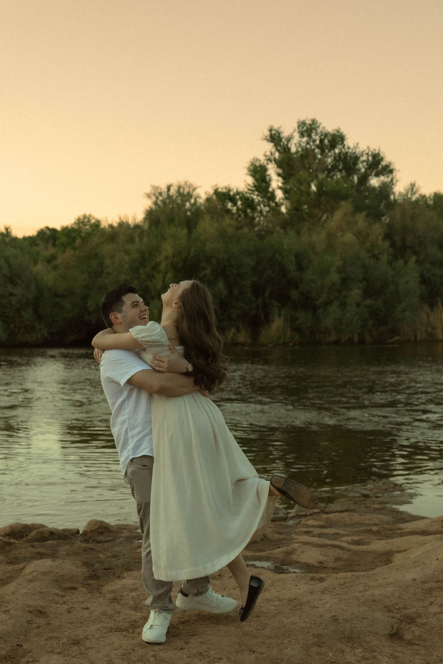 Man lifting woman in white dress on sandy riverbank at sunset with trees in background.