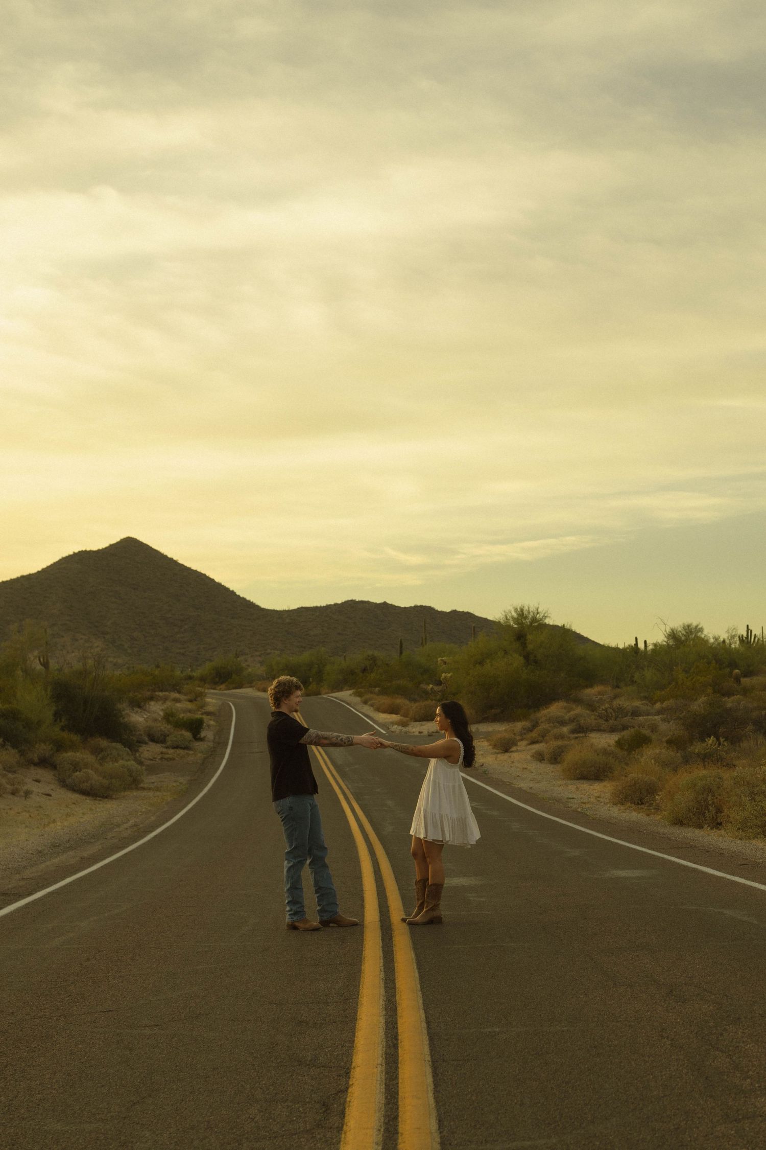 Couple holding hands on desert road centerline at golden hour with mountain backdrop.