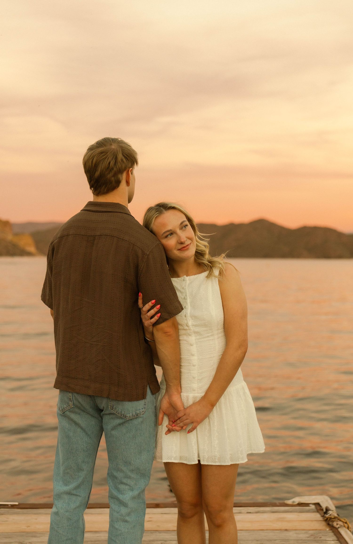 A couple embraces on a lakeside pier during golden hour with mountains in the background.