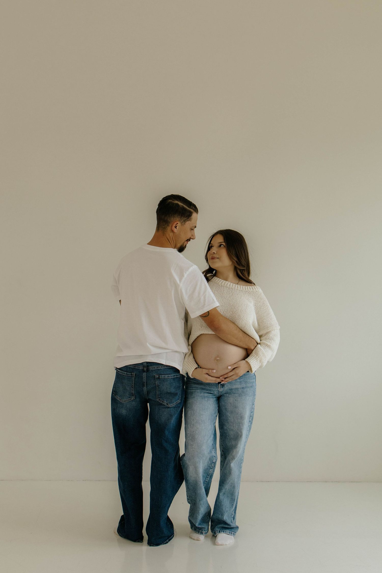 Two people in casual denim and white clothing sit close together against a minimalist white wall background.