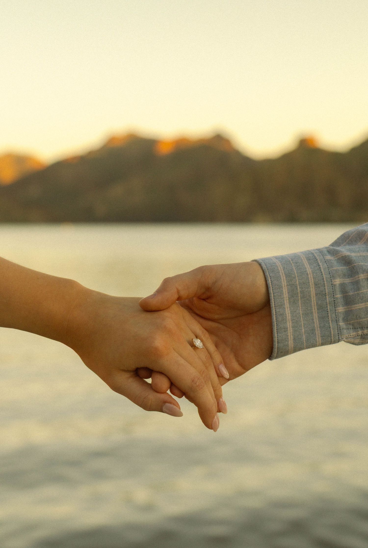 Two hands reaching out to hold each other against a sunset waterfront backdrop.
