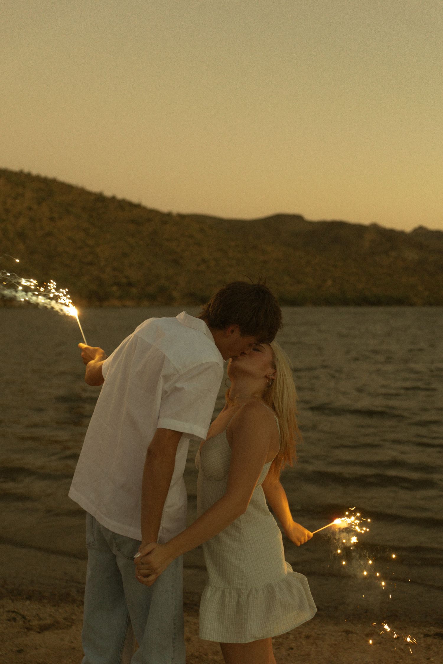Couple kissing on beach at sunset holding sparklers, with mountain lake backdrop in golden hour light.