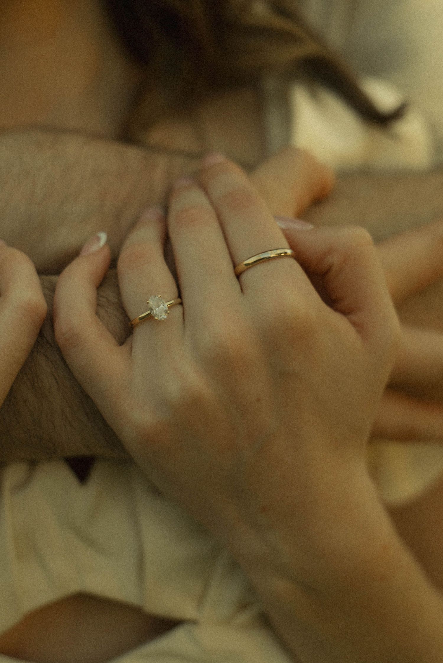 Close-up of womans hands wearing a diamond engagement ring and gold band resting on partners arm.