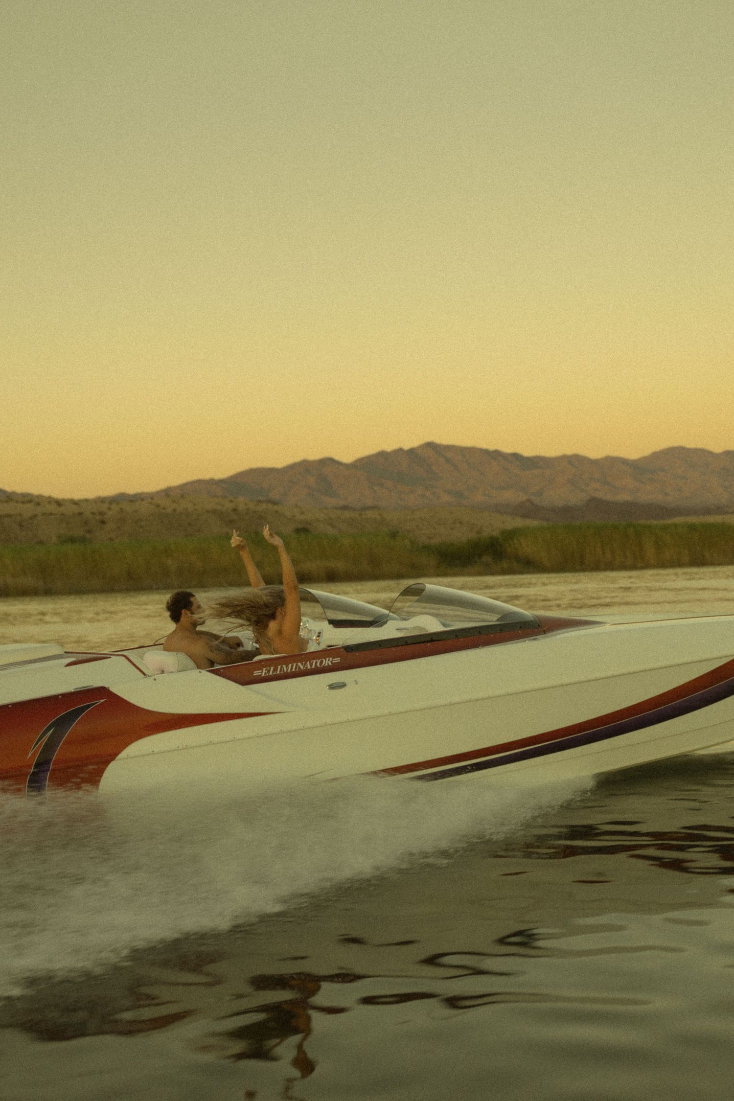 A couple rides a red and white speedboat across a lake with mountain backdrop at golden sunset.