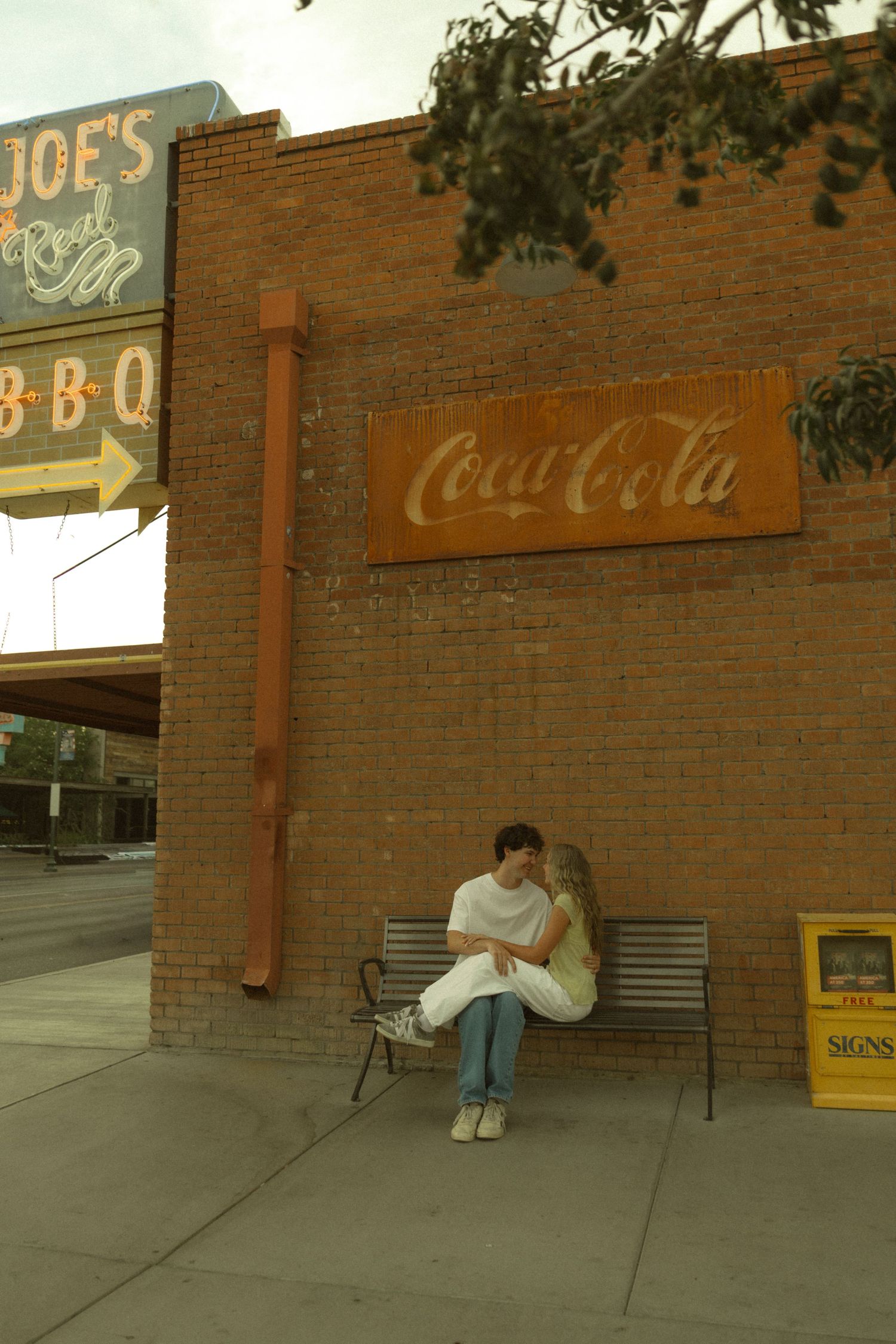 Romantic couple sitting on a bench outside a brick building with a vintage Coca-Cola sign.