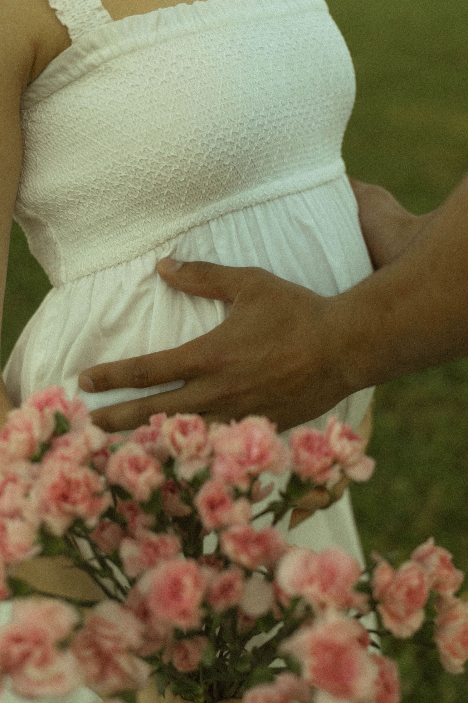 Partner gently holding pregnant woman's baby bump with pink carnations in foreground during maternity shoot.