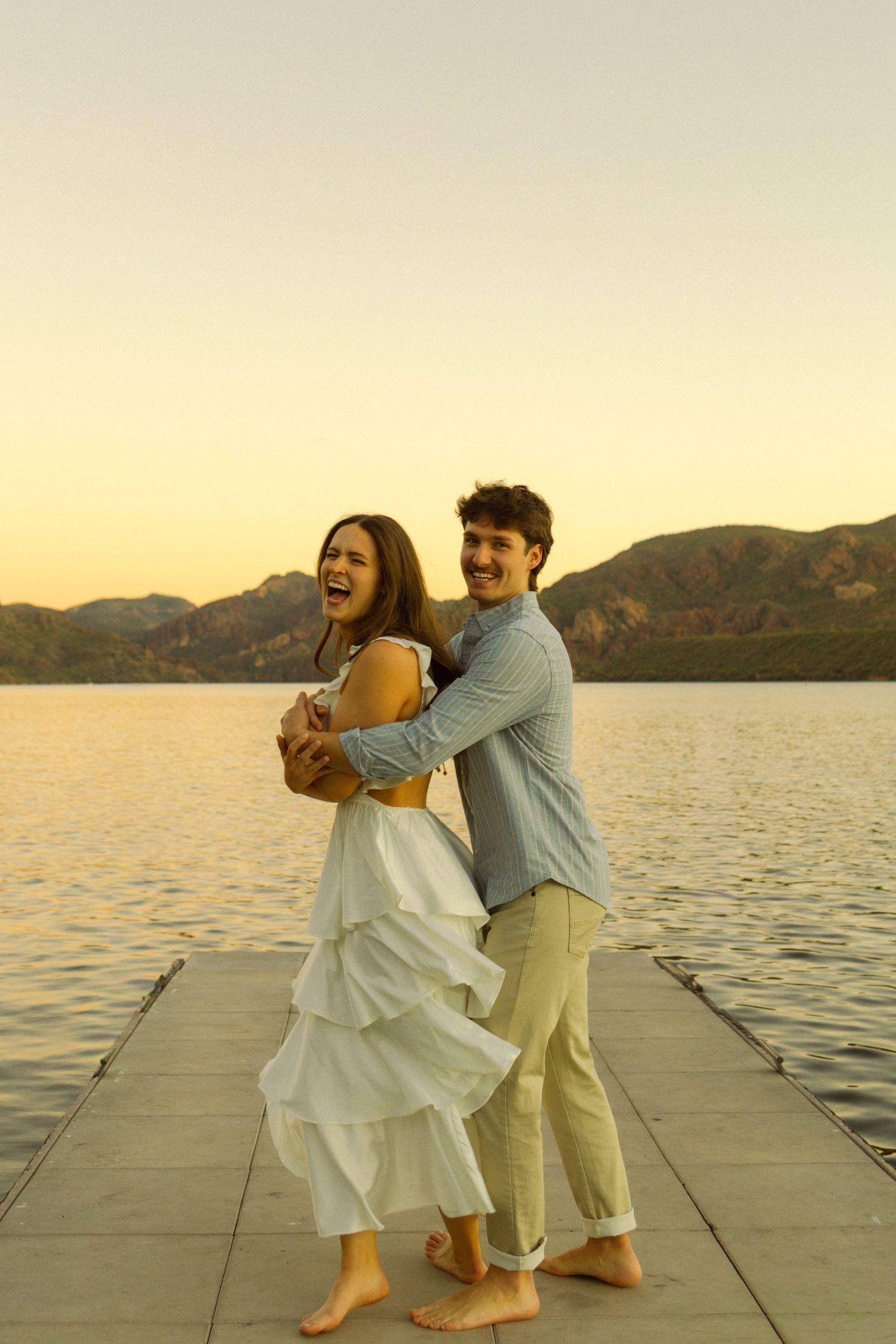 Joyful couple dancing barefoot on a dock at sunset with mountains in the background.
