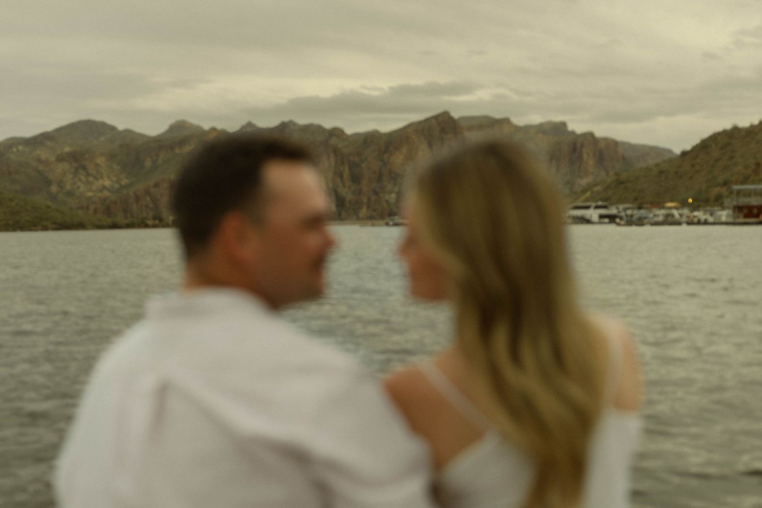 A couple stands together on a dock overlooking a serene mountain lake at sunset.