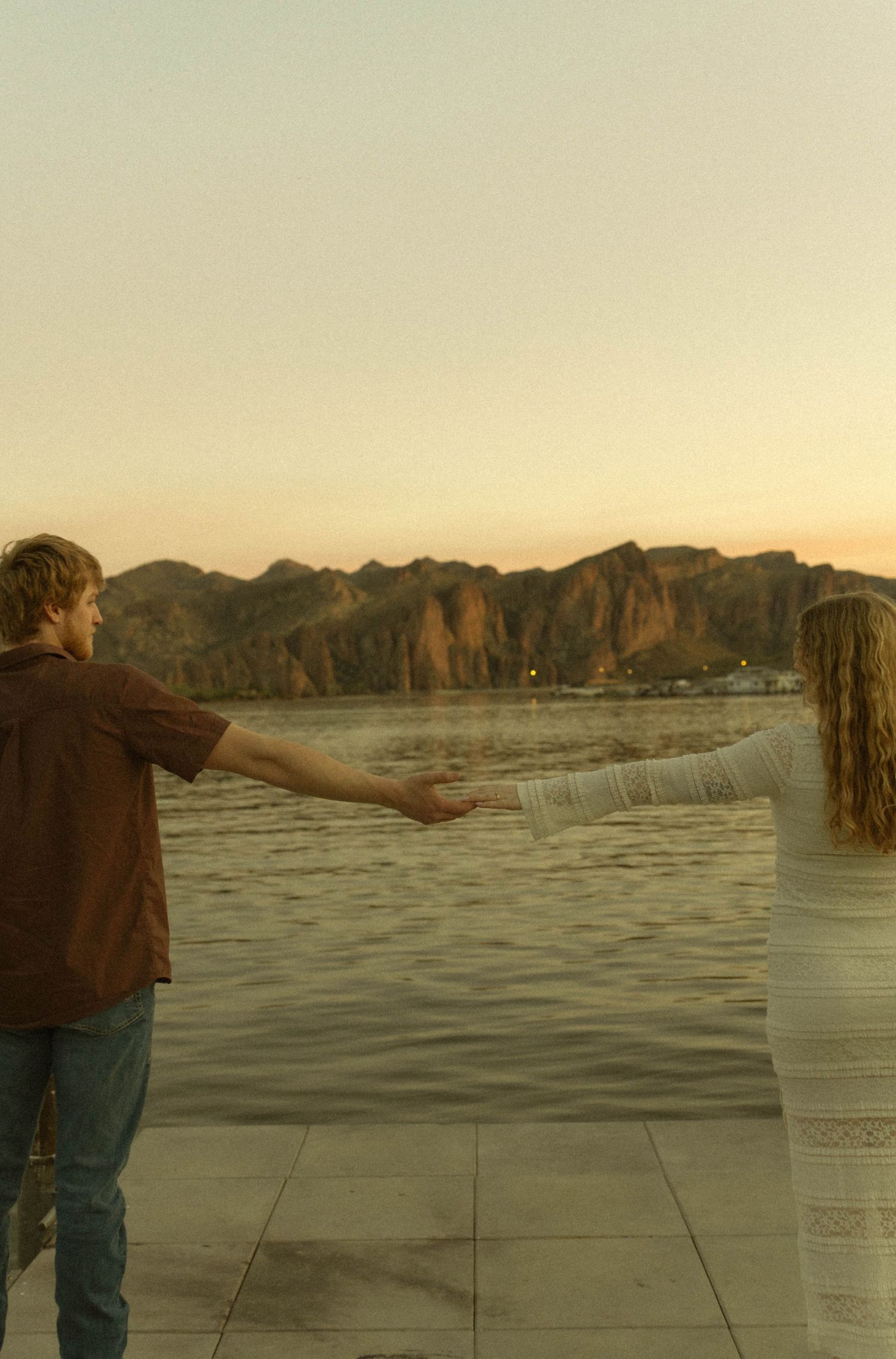 A person stands alone on a concrete surface with mountains and water in the background at sunset.