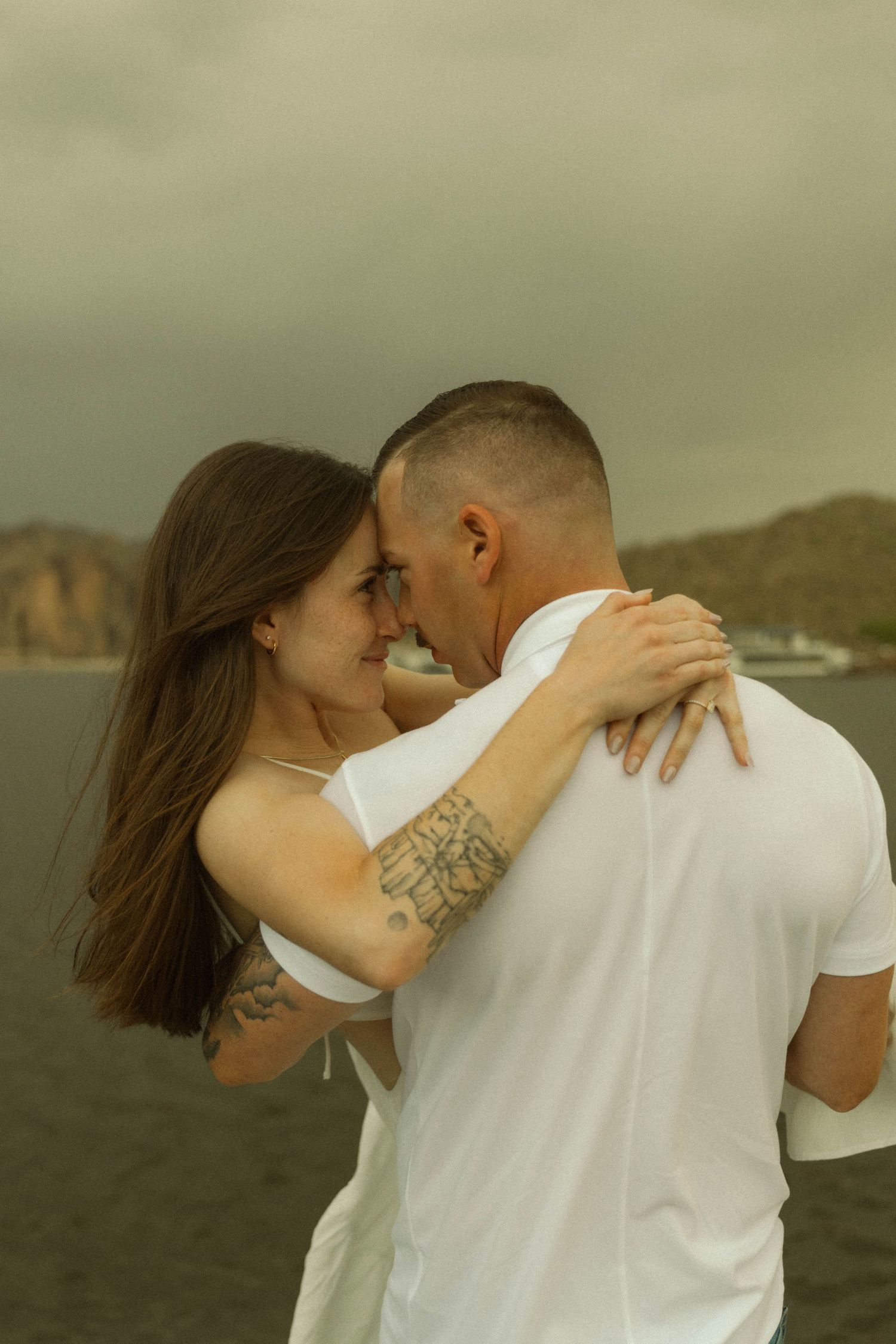 Intimate embrace between a couple with mountains and water in the background.