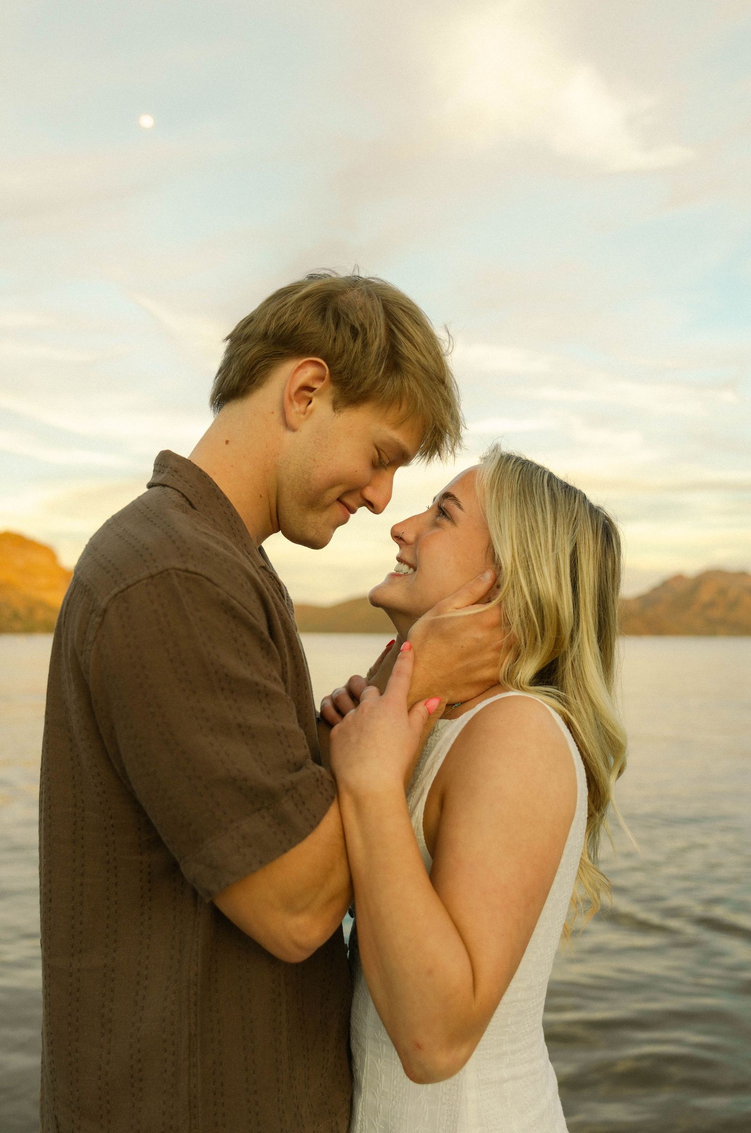Romantic couple embraces at sunset by a lake with mountains in the background, creating a tender moment.