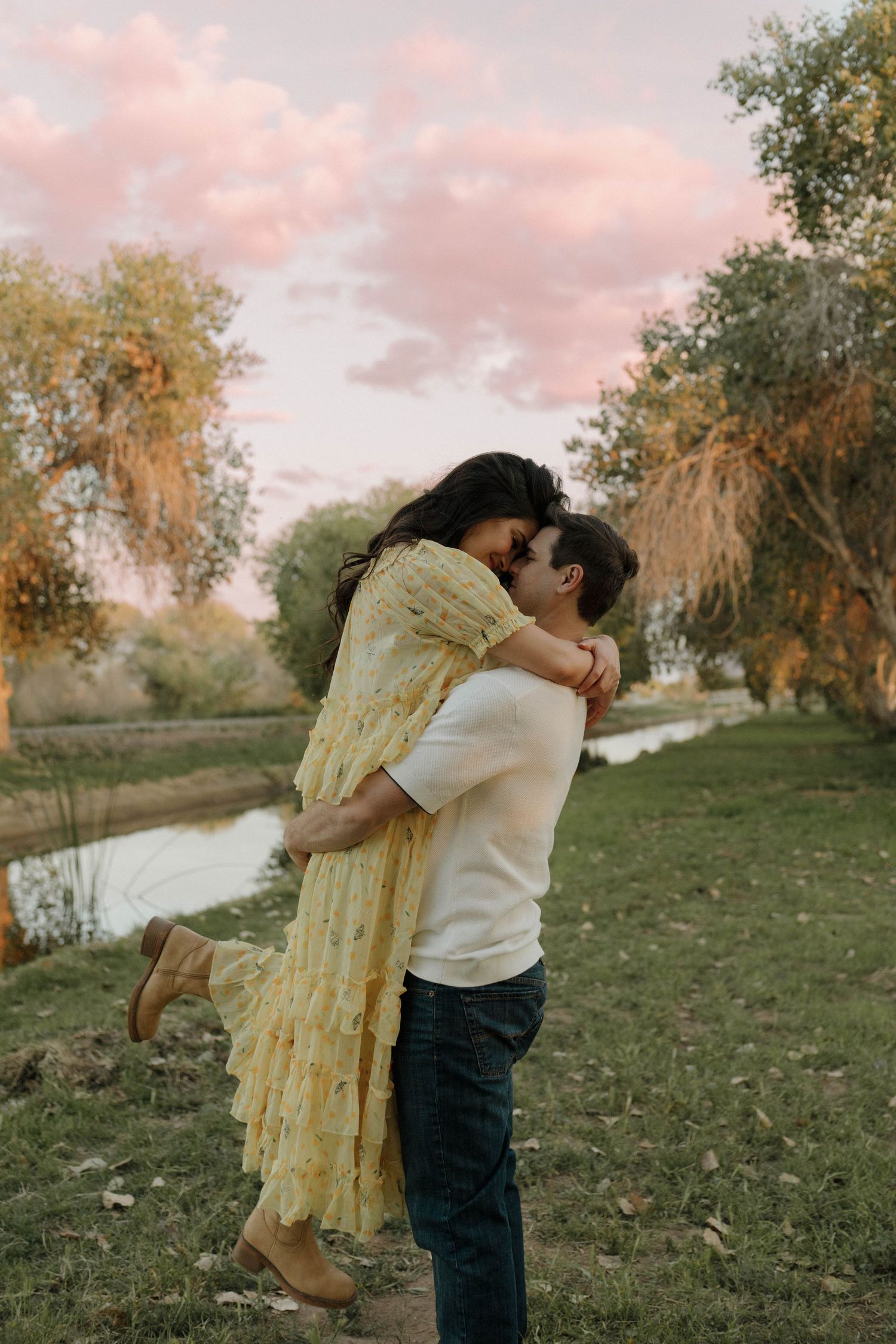 Romantic lift pose in yellow dress against sunset sky and autumn trees.