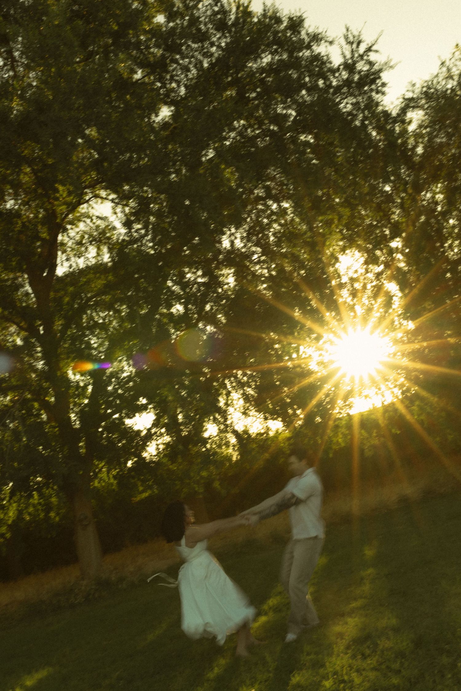Couple dancing in golden sunset light through green trees in a park, with beautiful sun flare and lens bokeh effect.