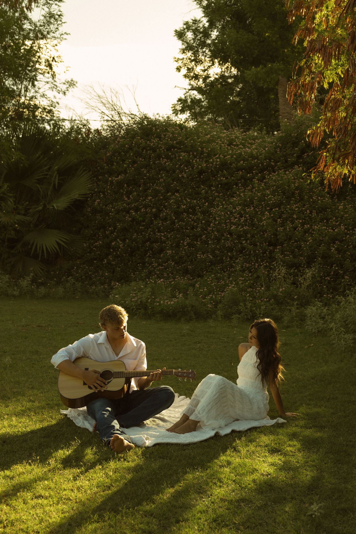 Couple enjoying a romantic sunset picnic on grass, man playing acoustic guitar for woman in white dress.