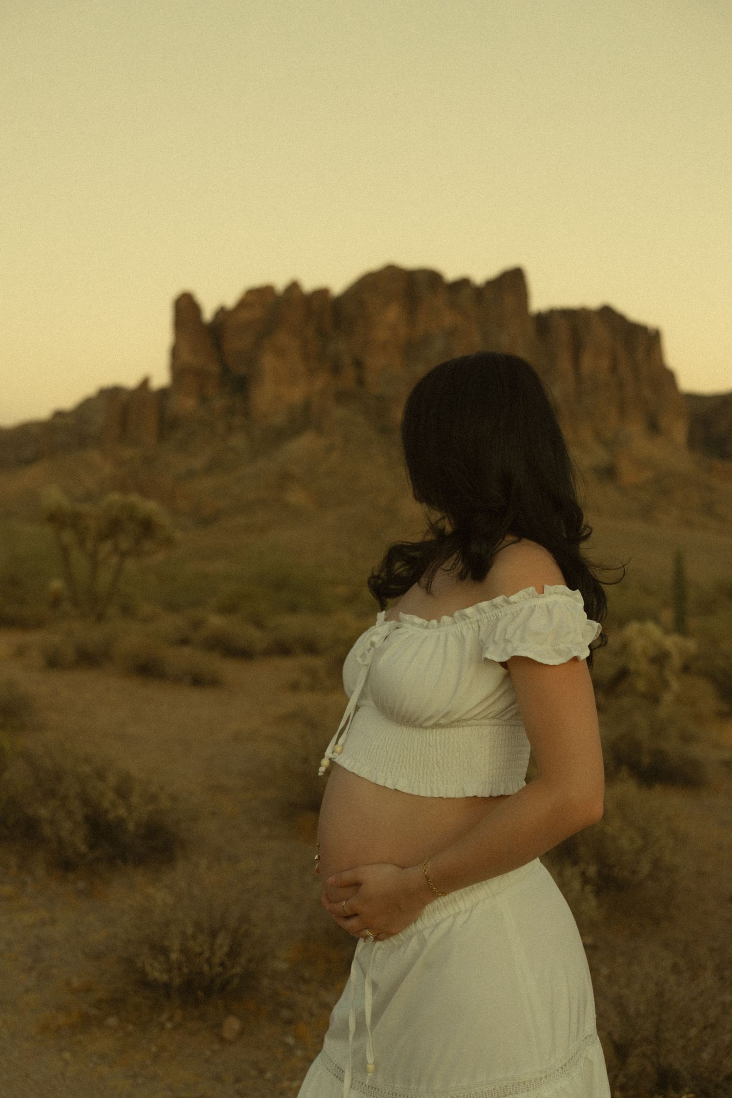 Pregnant woman in white off-shoulder dress cradling baby bump at desert sunset with rocky mountain backdrop.