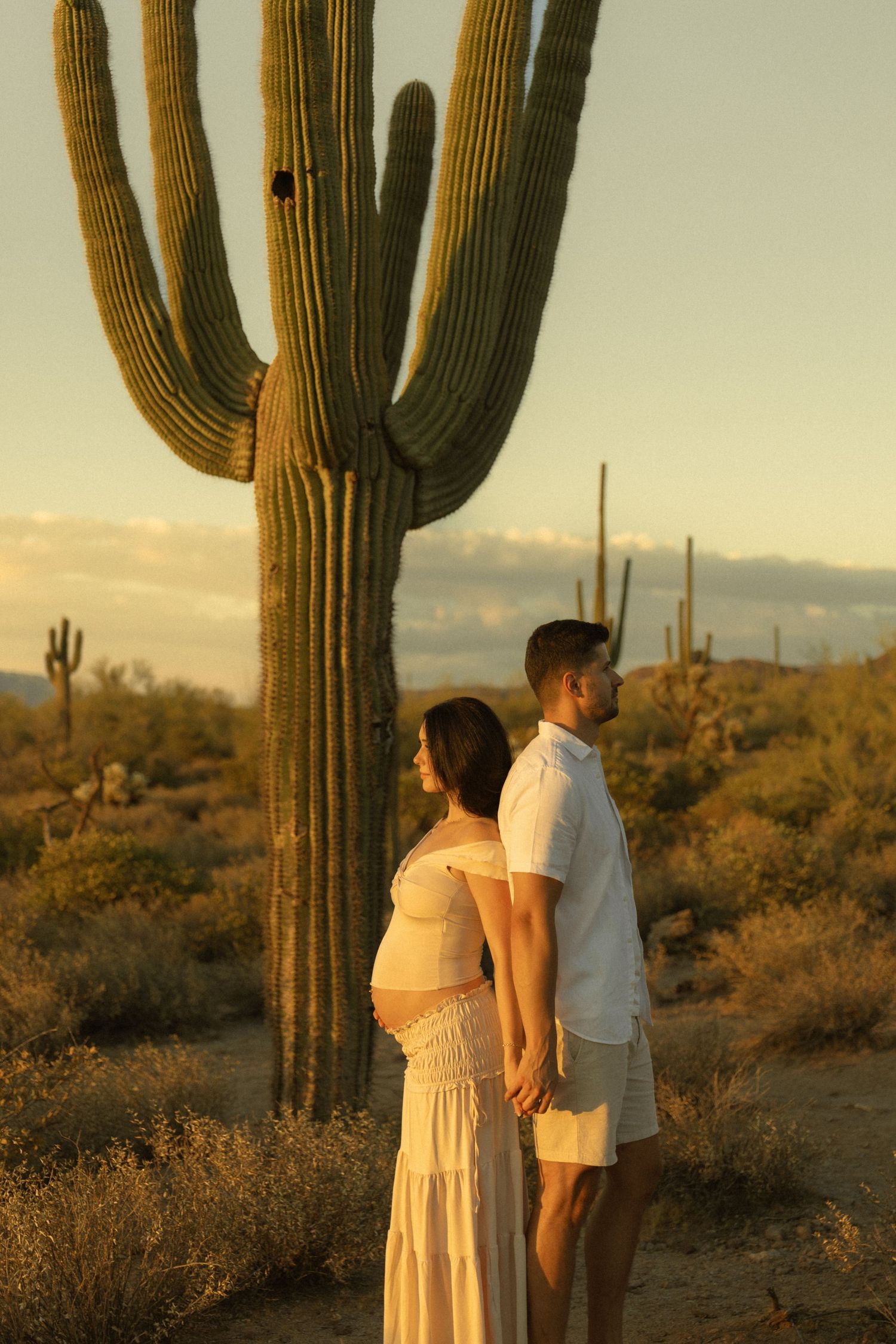 Pregnant couple holding hands in Arizona desert at golden hour with tall saguaro cactus backdrop.