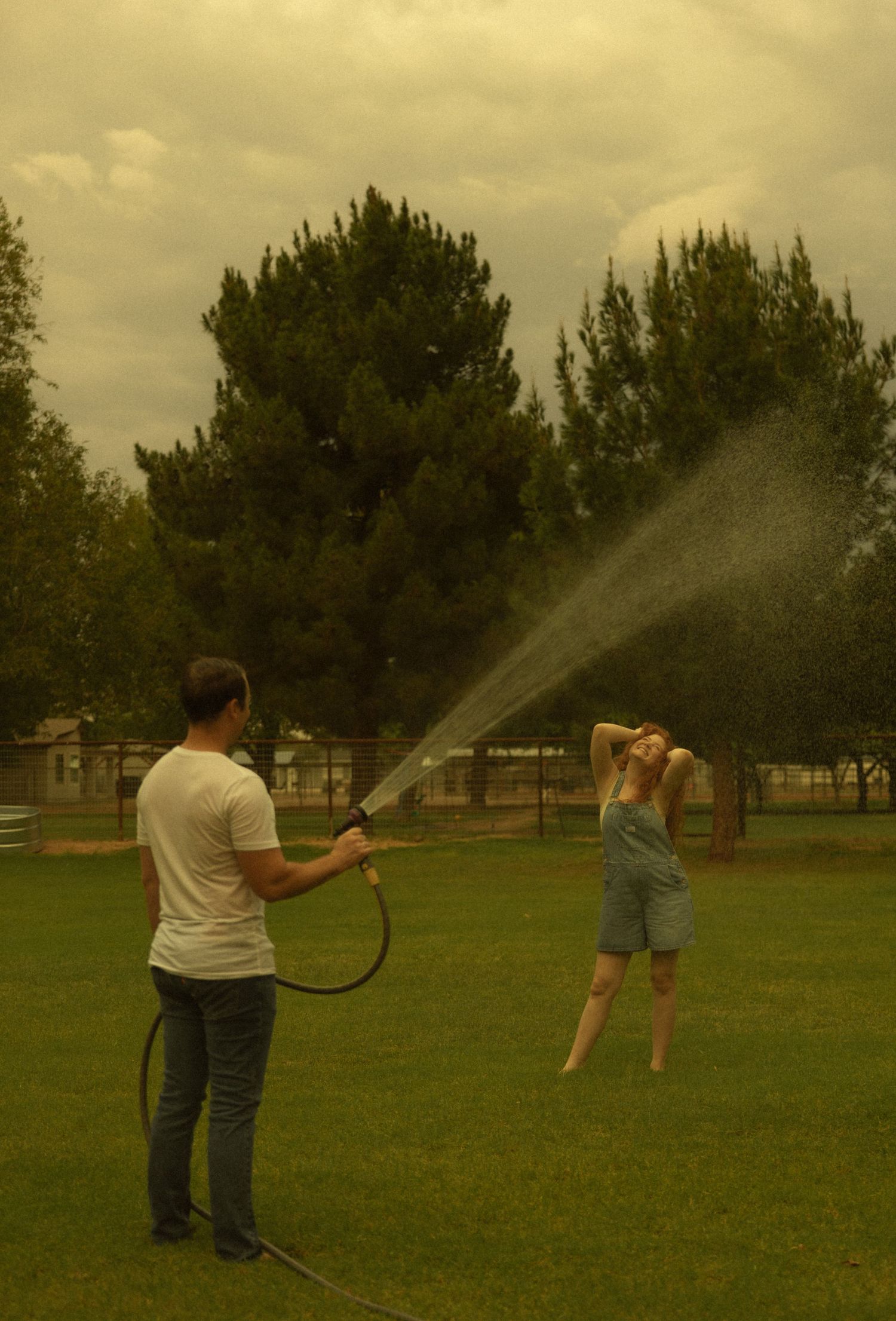 Man spraying woman with garden hose on green lawn with trees in background on cloudy day.