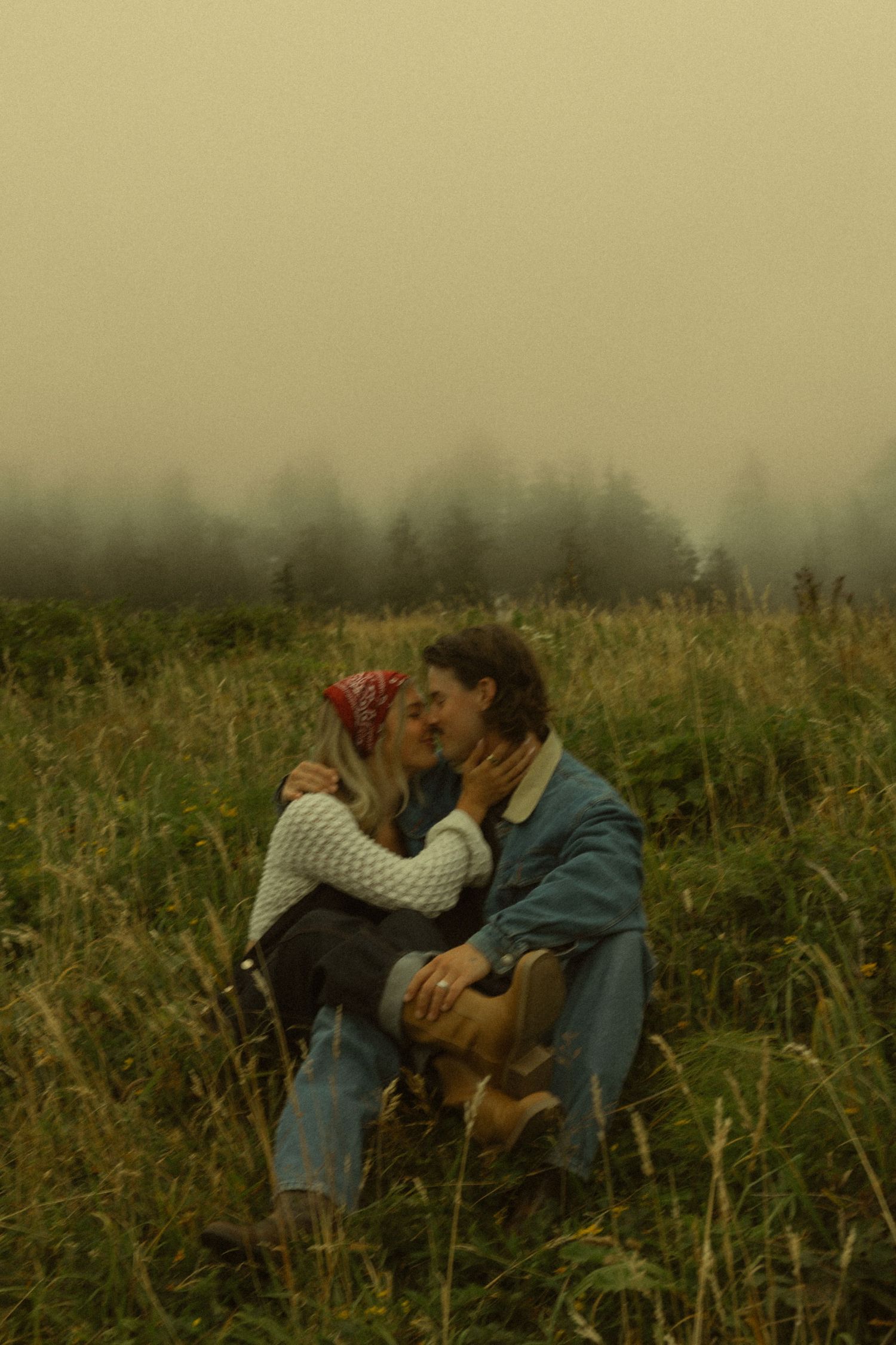 Couple sharing a romantic kiss in a misty meadow with tall grass and foggy pine trees in the background.
