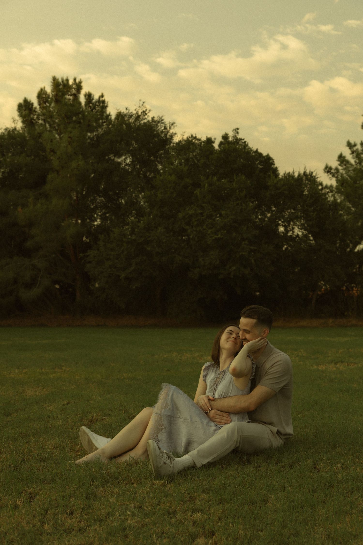 Couple sitting together on green grass in a park, embracing tenderly at sunset with trees in the background.