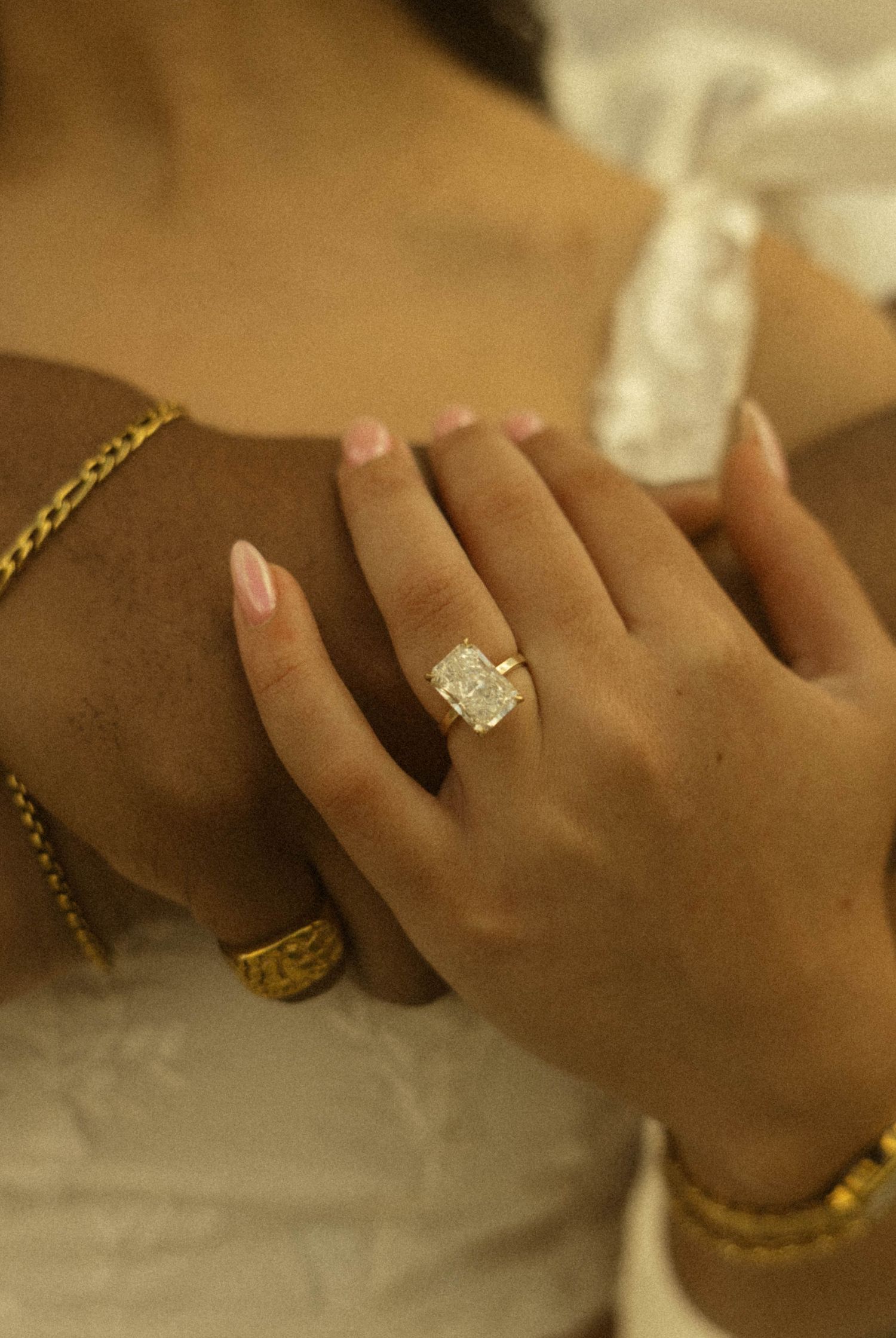Close-up of a woman's hand wearing a large radiant-cut diamond ring in gold setting, with pink nails and gold bracelets.