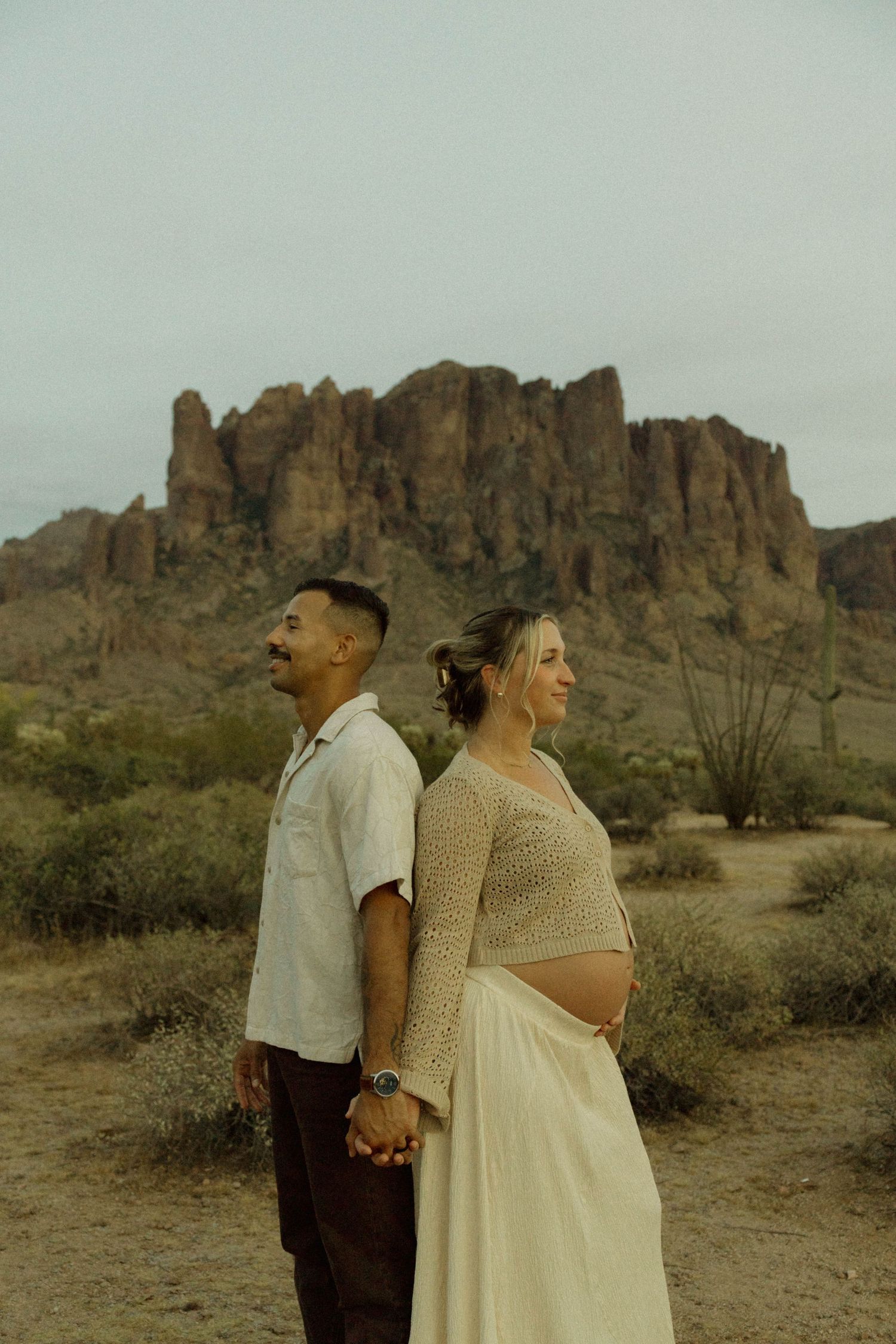 Pregnant couple holding hands back-to-back in Arizona desert with Superstition Mountains backdrop.