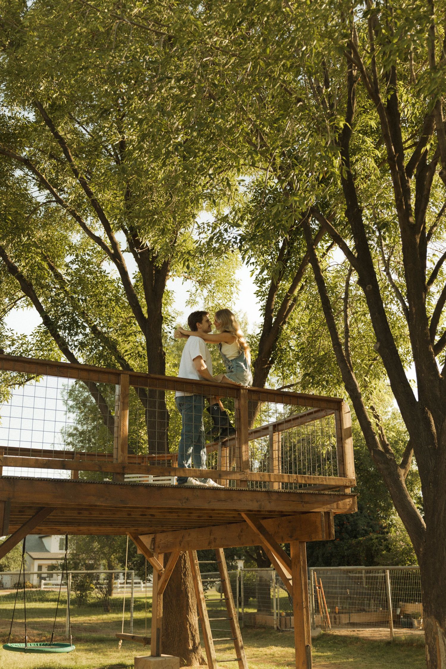 Couple sharing a romantic kiss on a wooden treehouse platform surrounded by lush green trees.
