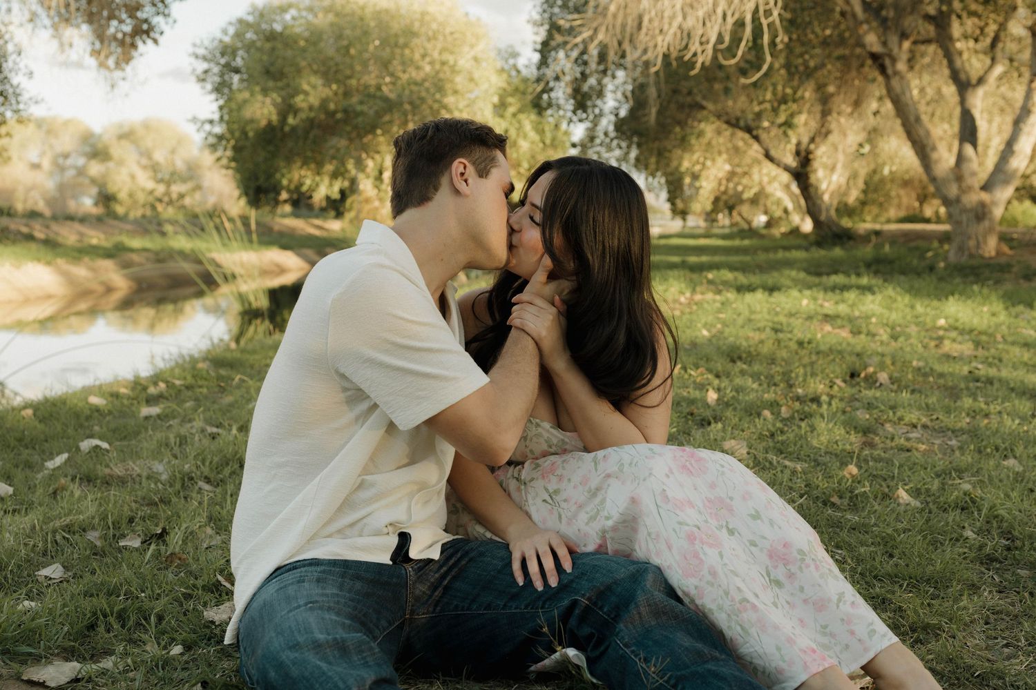 Couple embraces intimately while sitting in park surrounded by trees during sunset.