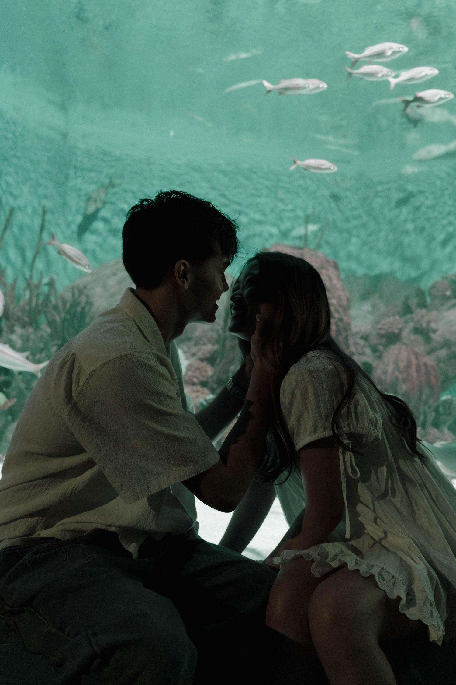 A romantic couple shares an intimate moment in front of an aquarium tank with fish swimming in turquoise waters.