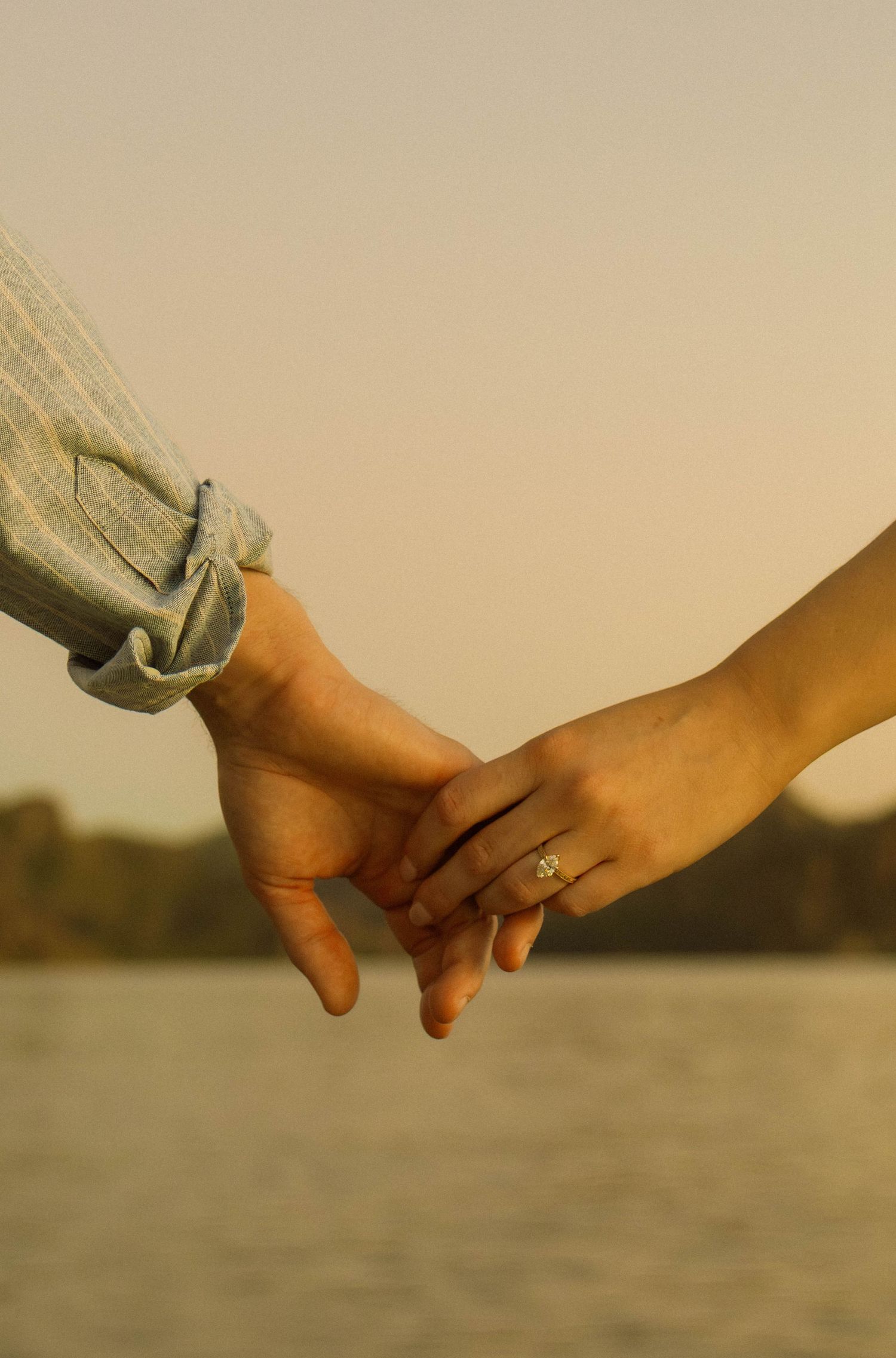 Close-up of hands holding with engagement ring visible against sunset lake backdrop.