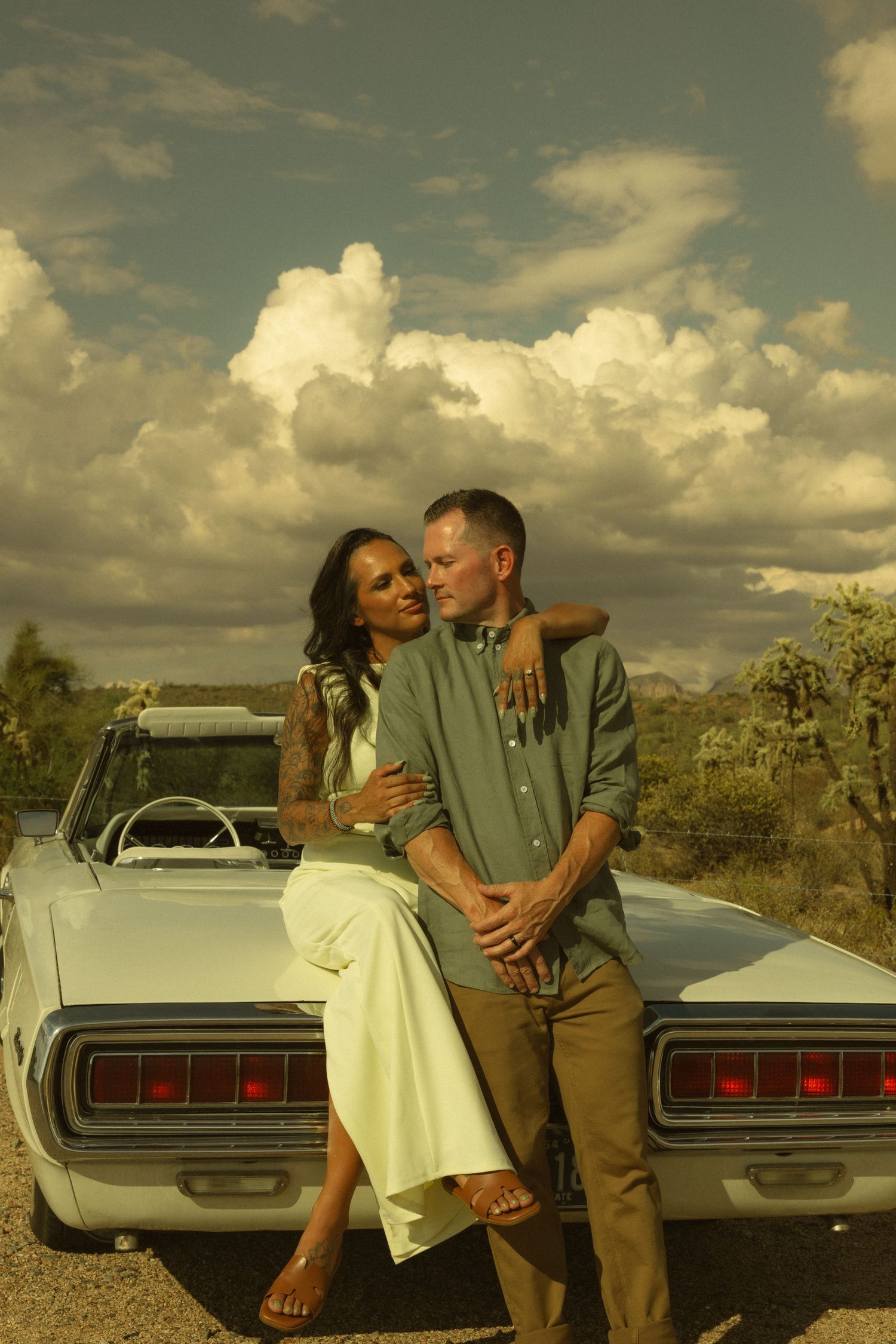 Couple embracing against a vintage convertible car in a desert landscape with dramatic cloudy sky.