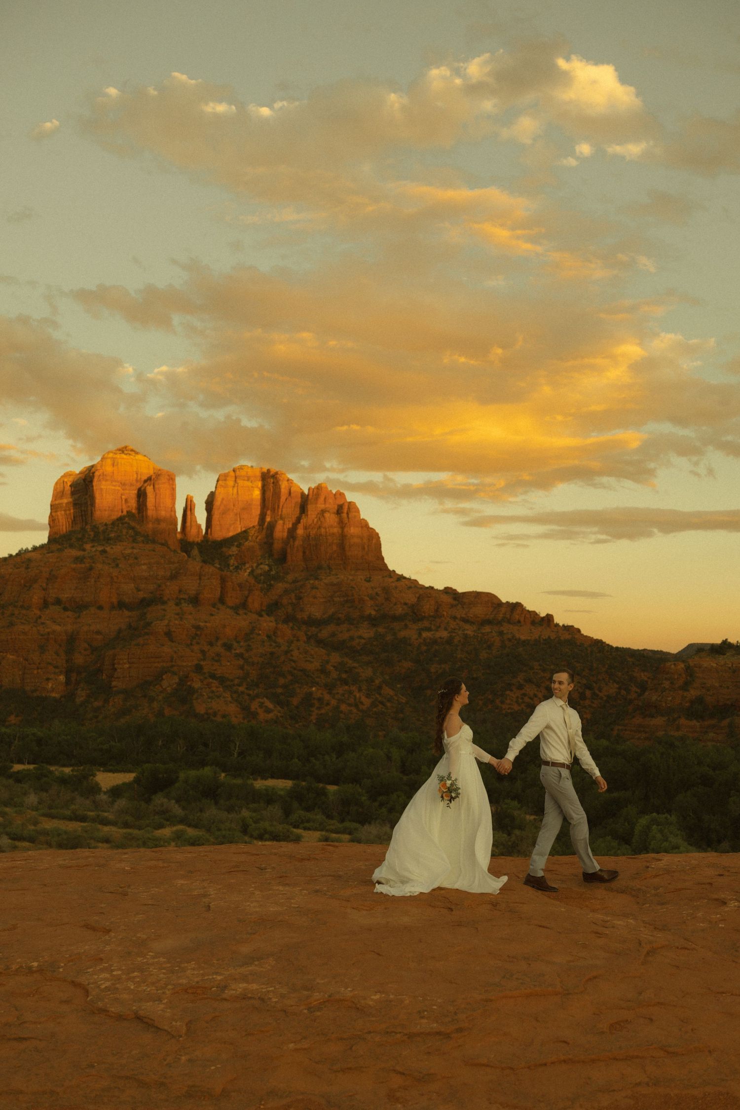 Bride and groom hold hands at sunset with Cathedral Rock in Sedona, Arizona glowing golden in the background.