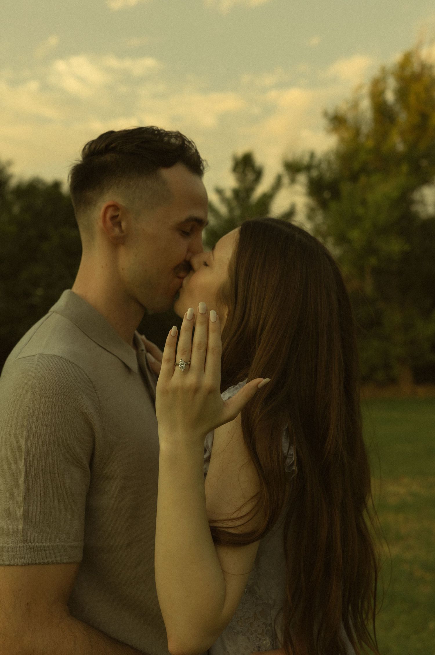 A couple shares a romantic almost-kiss outdoors, with the woman's hand gently touching his face, wearing an engagement ring.