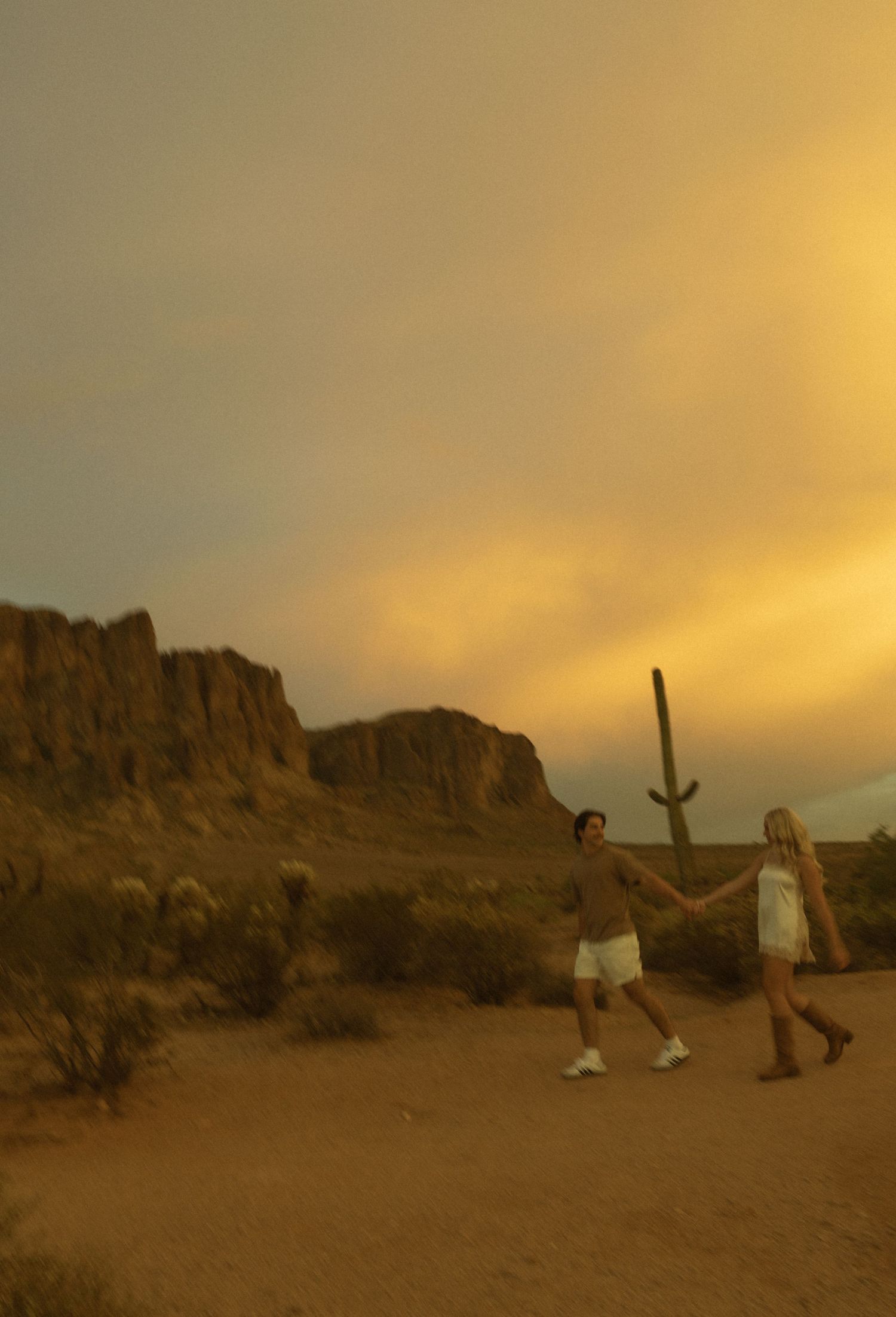 Couple walking hand in hand through Arizona desert with dramatic rocky mountains at sunset.