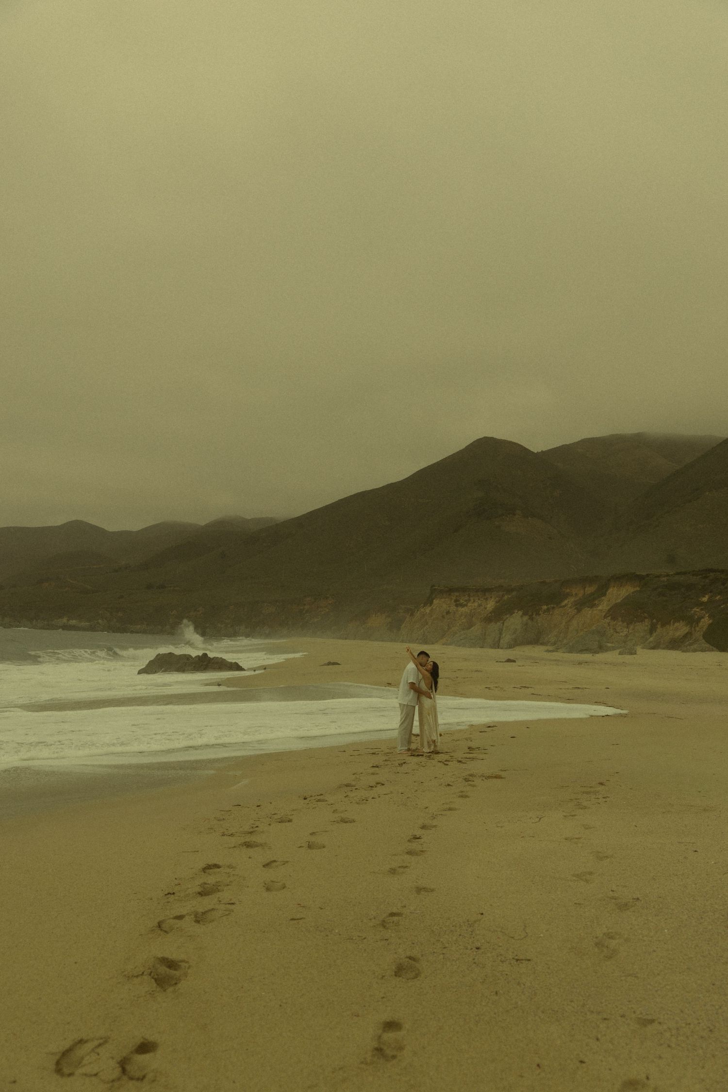 Romantic couple embracing on misty beach shoreline with dramatic coastal cliffs and ocean waves in background.