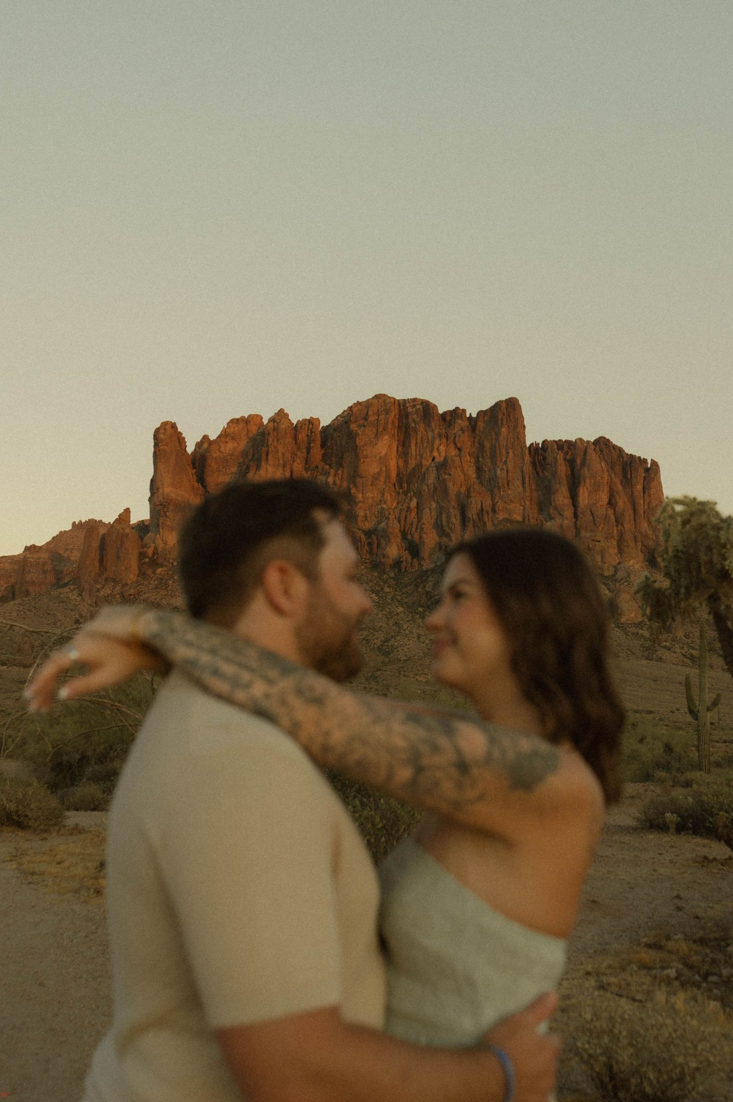 Couple embracing at sunset with dramatic red rock formations of the Superstition Mountains behind them.