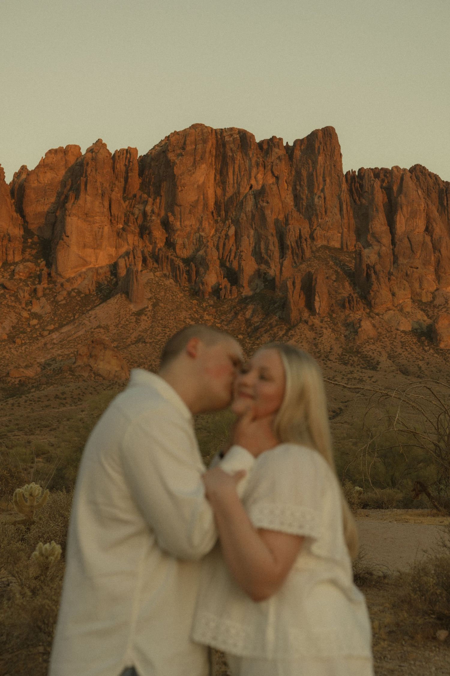 Couple sharing a romantic kiss at golden hour with stunning red rock Superstition Mountains in the Arizona desert background.