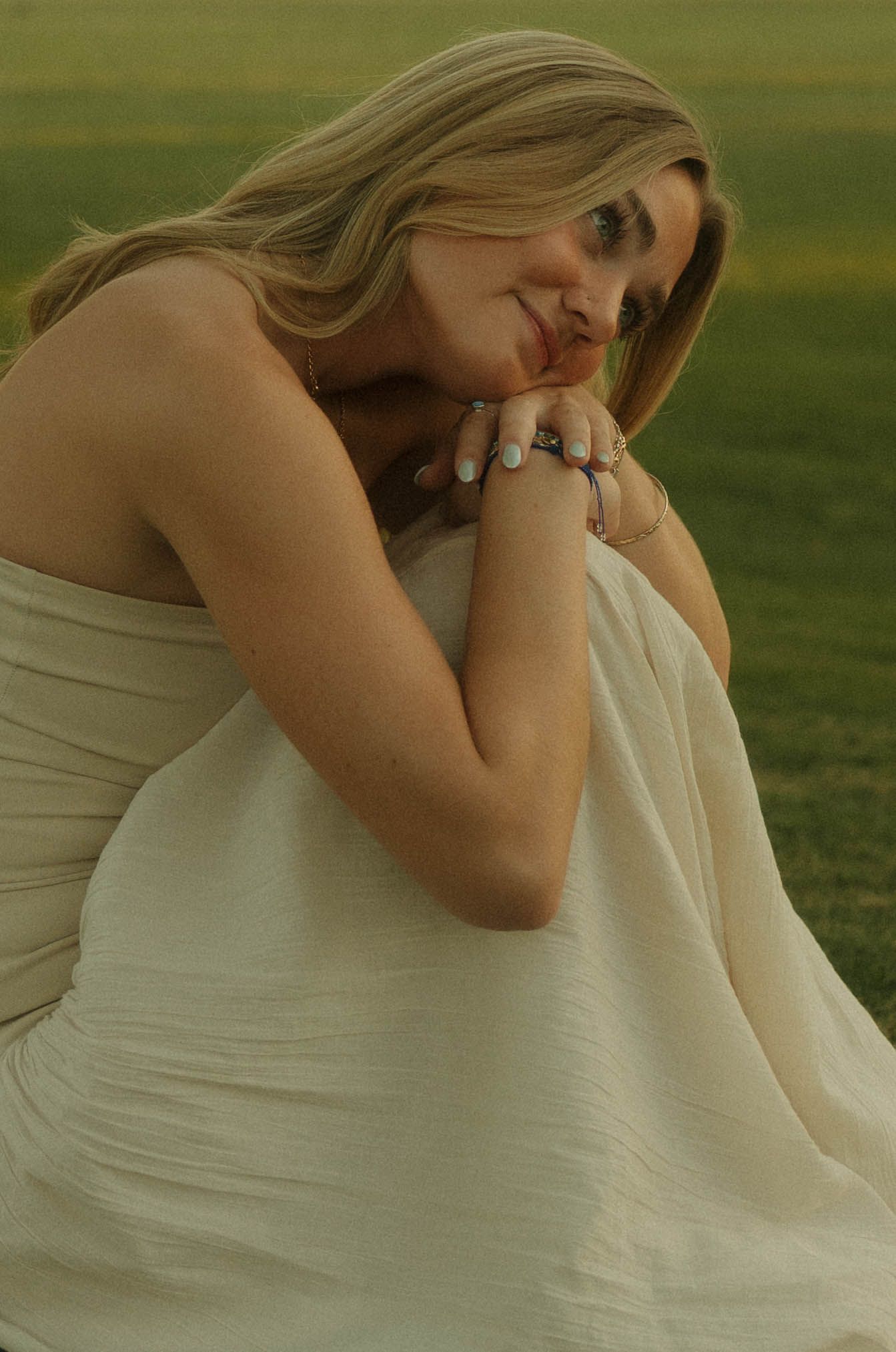 A blonde woman in a cream strapless dress sits gracefully outdoors, resting her chin on her hands with rings on her fingers.