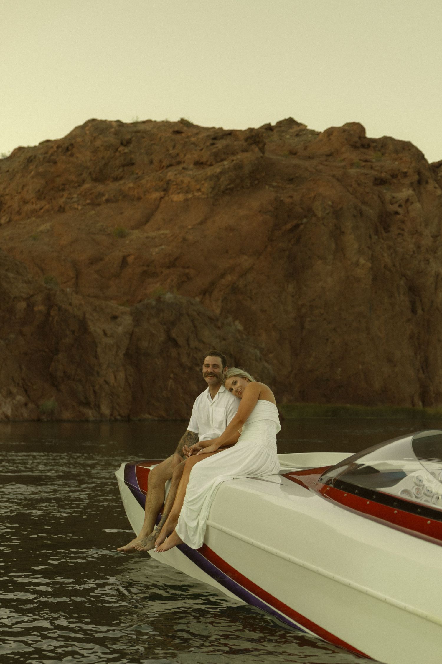 Couple in white sitting on bow of speedboat with red rock cliffs backdrop on calm water.