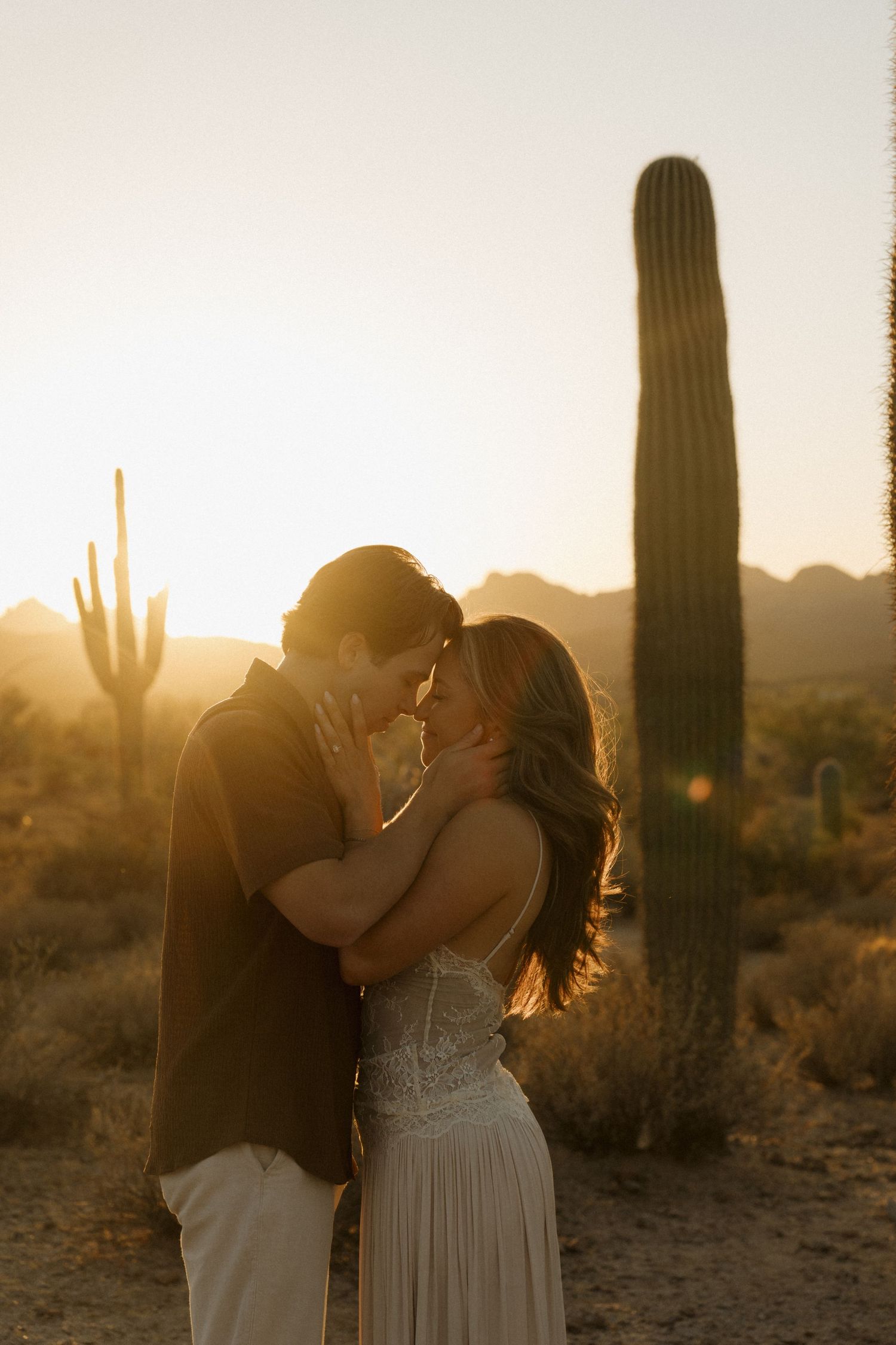 Couple sharing a romantic kiss at golden hour in the Arizona desert with saguaro cacti silhouetted behind them.